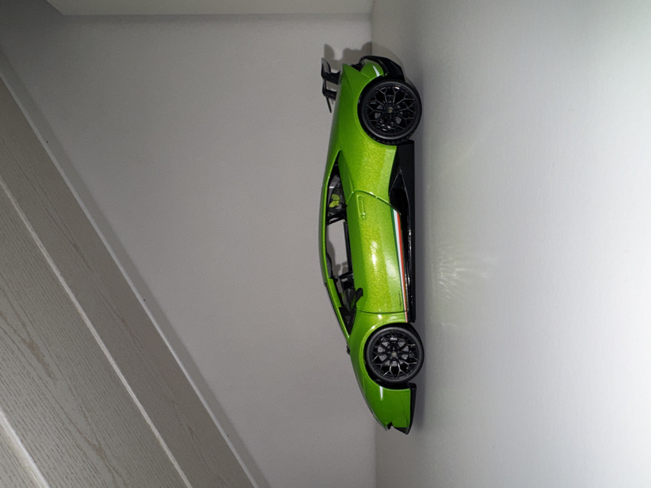 A lime green toy car is displayed vertically on a white wall next to a staircase. The car has black wheels, a black spoiler, and black trim. A green, white, and red stripe is visible along the car's side. The car's details are crisp and show the intricacies of the model.