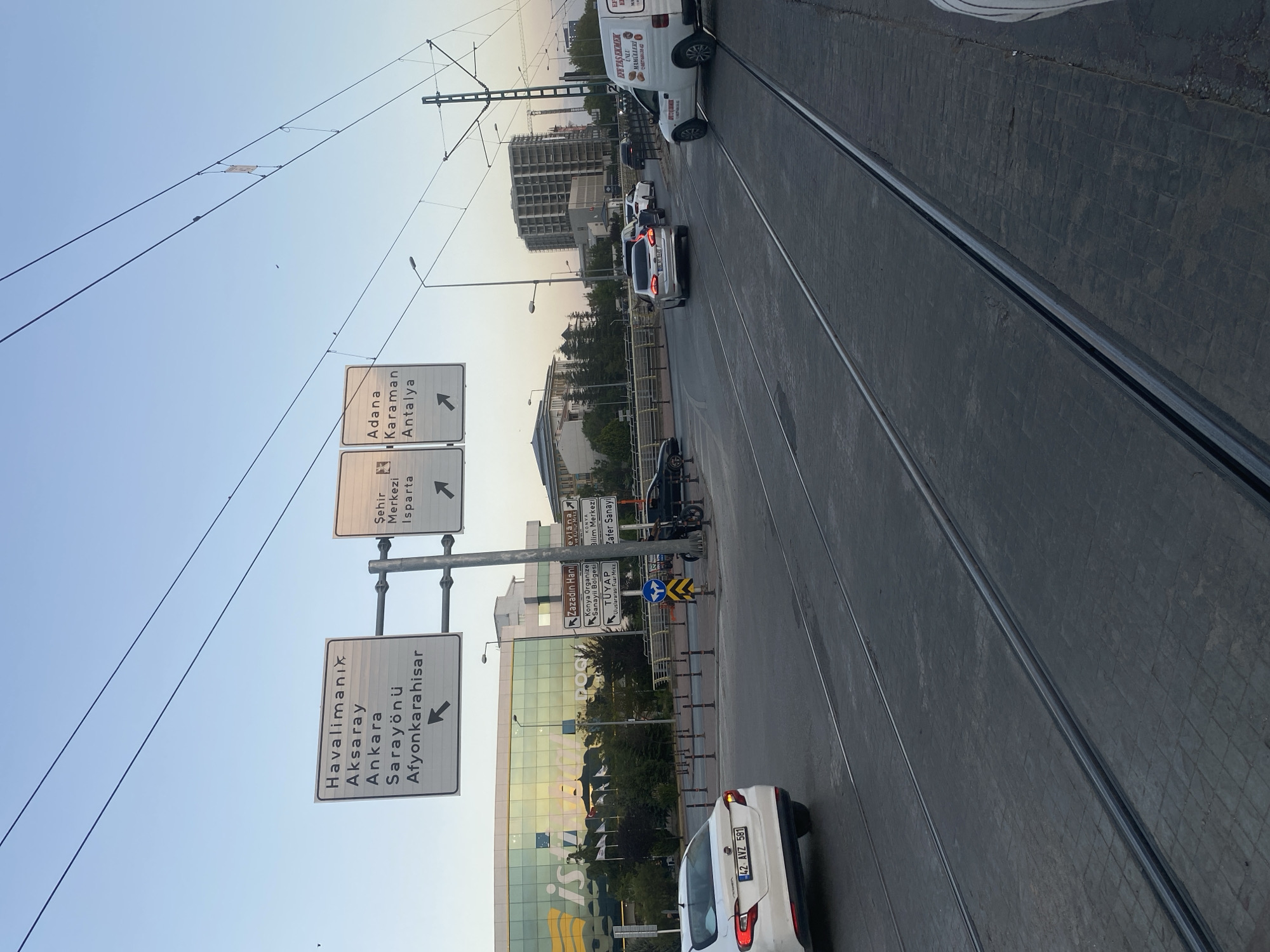 The image depicts a street scene in Konya, Turkey, featuring a junction with multiple directional signs. In the foreground, tram tracks run along a paved road. Several vehicles are visible, including a white car in the left lane with the license plate "42 AVZ 581," a silver sedan in the right lane, and other cars further down the road. A white van with text on its side is also present.
Dominating the background is a large, modern building with a reflective glass facade, likely a commercial or institutional building, bearing the partial word "ist" which may be part of "İstanbul." To the left of this building, a street sign indicates directions to "Havalimanı" (Airport), "Aksaray," "Ankara," "Sarayönü," and "Afyonkarahisar." To the right, another sign points towards "Şehir Merkezi" (City Center) and "Isparta." Further down the road, additional signs indicate "Zazadin Hanı," "Konya Organize Sanayi Bölgesi," "TÜYAP Uluslararası Fuar Merkezi," "Evlana ve Kültür Merkezi," "Bilim Merkezi," and "Zafer Sanayi."
The sky is clear with a gradient of blue, suggesting it is late afternoon or early evening. Overhead, tram wires are strung between poles, and a street lamp is visible. The overall impression is of a busy urban intersection in Konya.