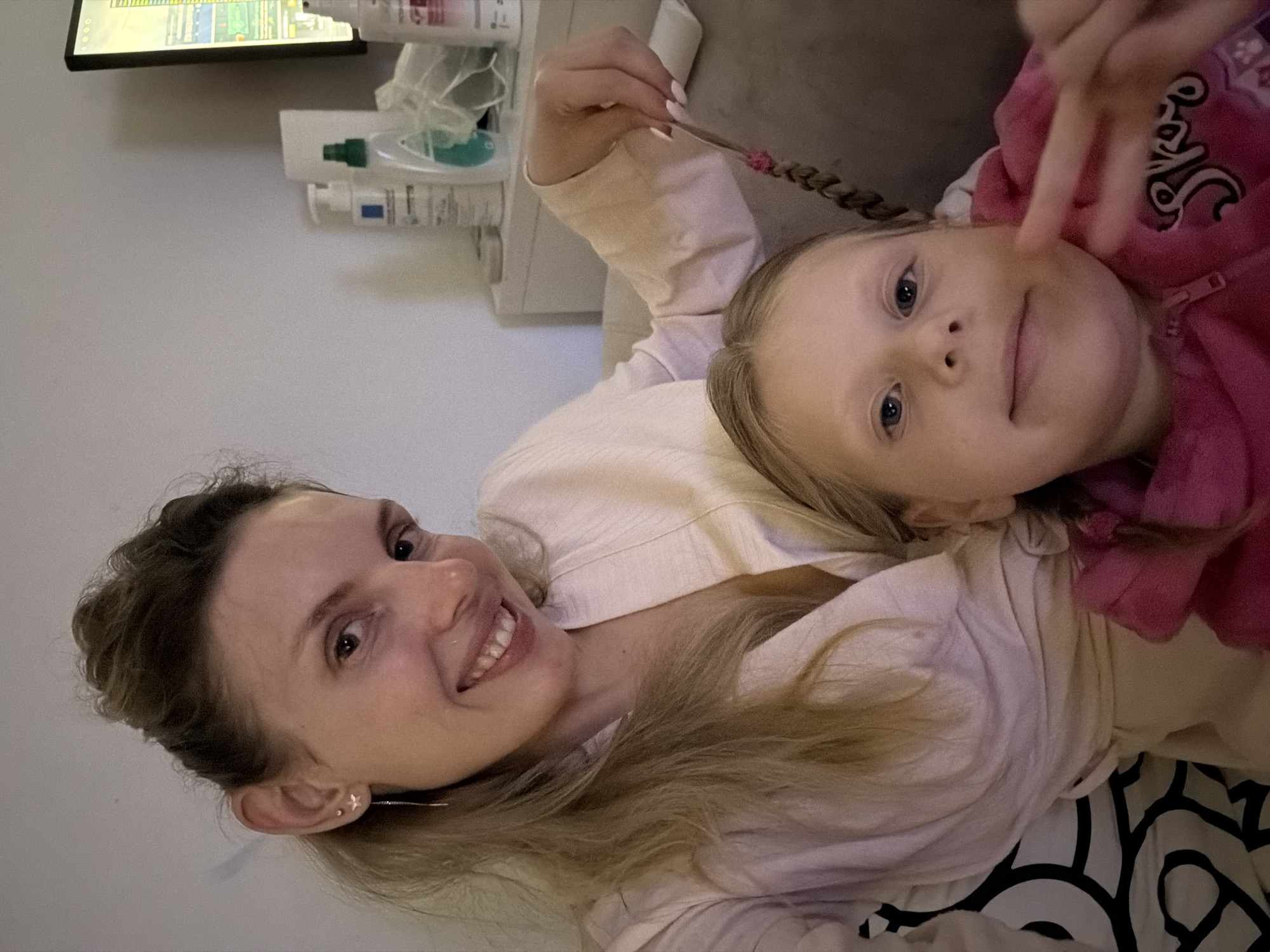 A woman and a young girl are lying down close to each other, facing the camera. The woman is on the left and smiling, with blonde hair. The girl is on the right, wearing a pink hooded top, and making a peace sign with her fingers. Her hair is blonde and braided. There is a sofa behind them. In the background, there is a shelf with various toiletries and a screen displaying data. The photo was likely taken indoors. There is no indication that this photo was taken in Budva, Montenegro.
