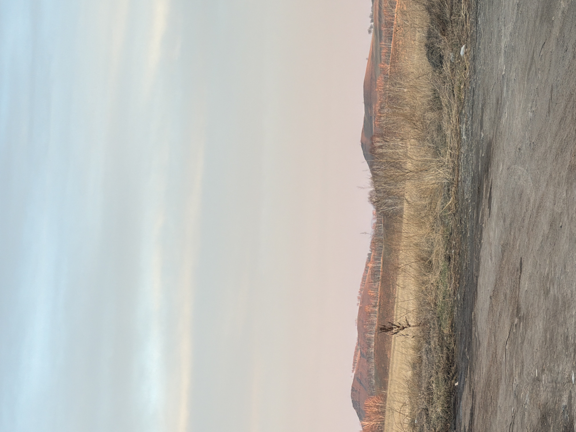A landscape photograph oriented vertically features a gradient of sky on the left, transitioning from a light blue at the top to a pale orange near the center. To the right, a hilly terrain with sparse vegetation is visible, interspersed with what appear to be trees. An unpaved, gray dirt road stretches along the bottom edge of the photograph. Bazandaikha, Russia.