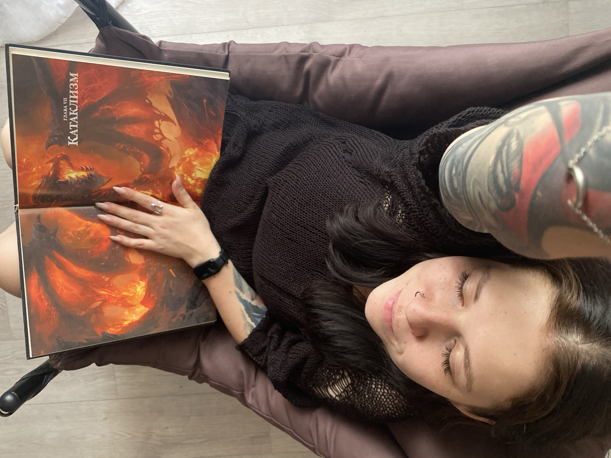 A woman is lying in a chair, holding a book with the title "КАТАКЛИЗМ" (Cataclysm) in Cyrillic script. The book's cover depicts a fiery scene with a dragon-like creature. The woman has long, dark hair, fair skin, a nose ring, and visible freckles. She is wearing a dark, textured sweater. Her left arm has a visible tattoo. Her nails are long and manicured. She is wearing a watch. The chair appears to be upholstered in a brown, cushioned fabric. The floor is visible in the background and seems to be light-colored. No location is identifiable in the image.