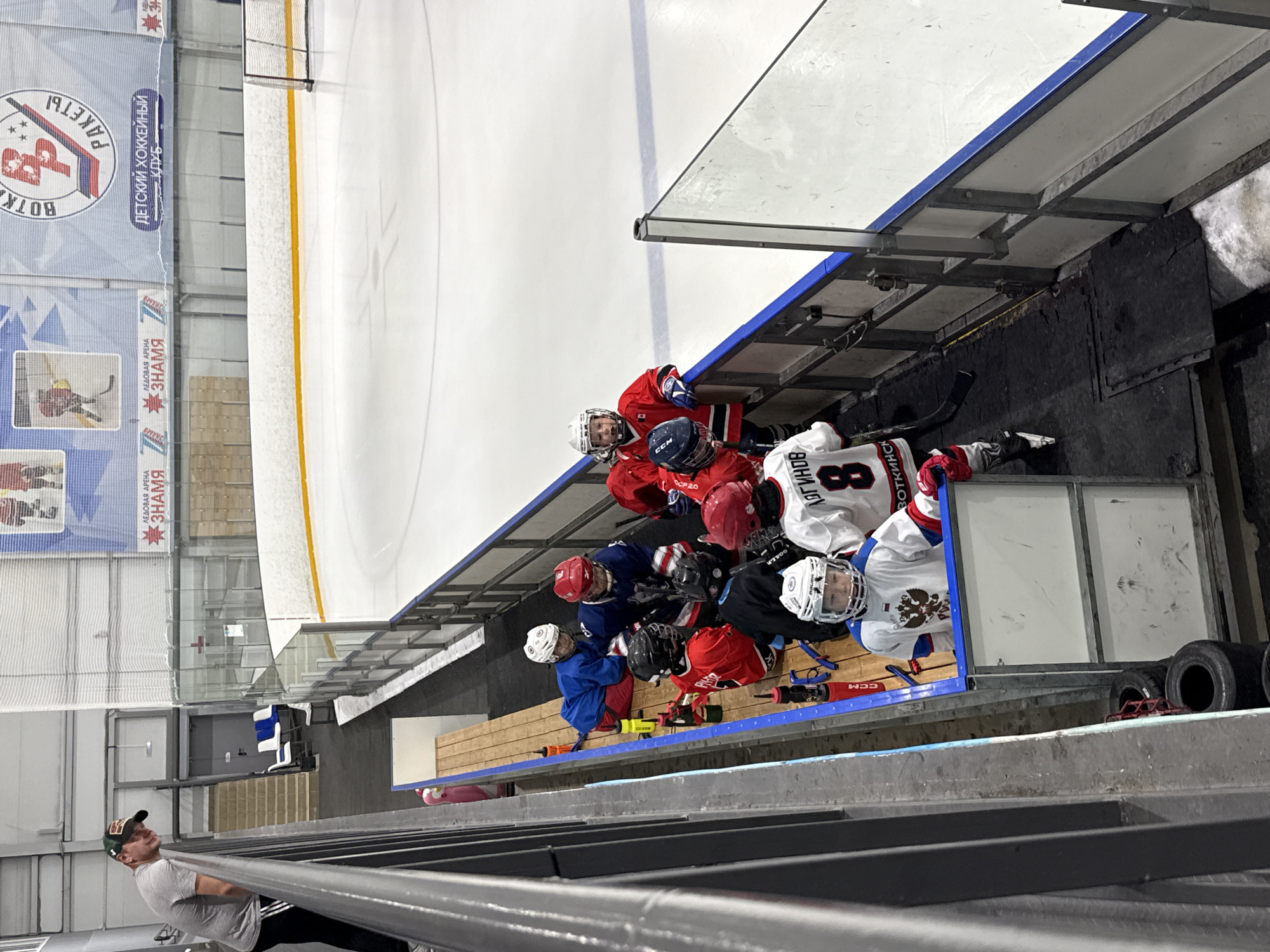 This is a photograph taken at an ice hockey arena in Votkinsk, Russia. The image features a group of young hockey players, most of whom are wearing red and white uniforms and helmets. They appear to be gathered on the bench, possibly during a break in a game or practice. The arena has an ice rink with markings and a yellow line, a blue-edged bench, and some equipment visible, including hockey sticks and what look like water bottles. In the background, banners with Russian text and logos related to "Votkinsk Rockets" and "Ice Arena Znamya" are displayed. A man in a grey t-shirt and a baseball cap is visible in the foreground, looking up. The lighting suggests it is daytime. The overall atmosphere appears to be focused on the youth hockey activity.