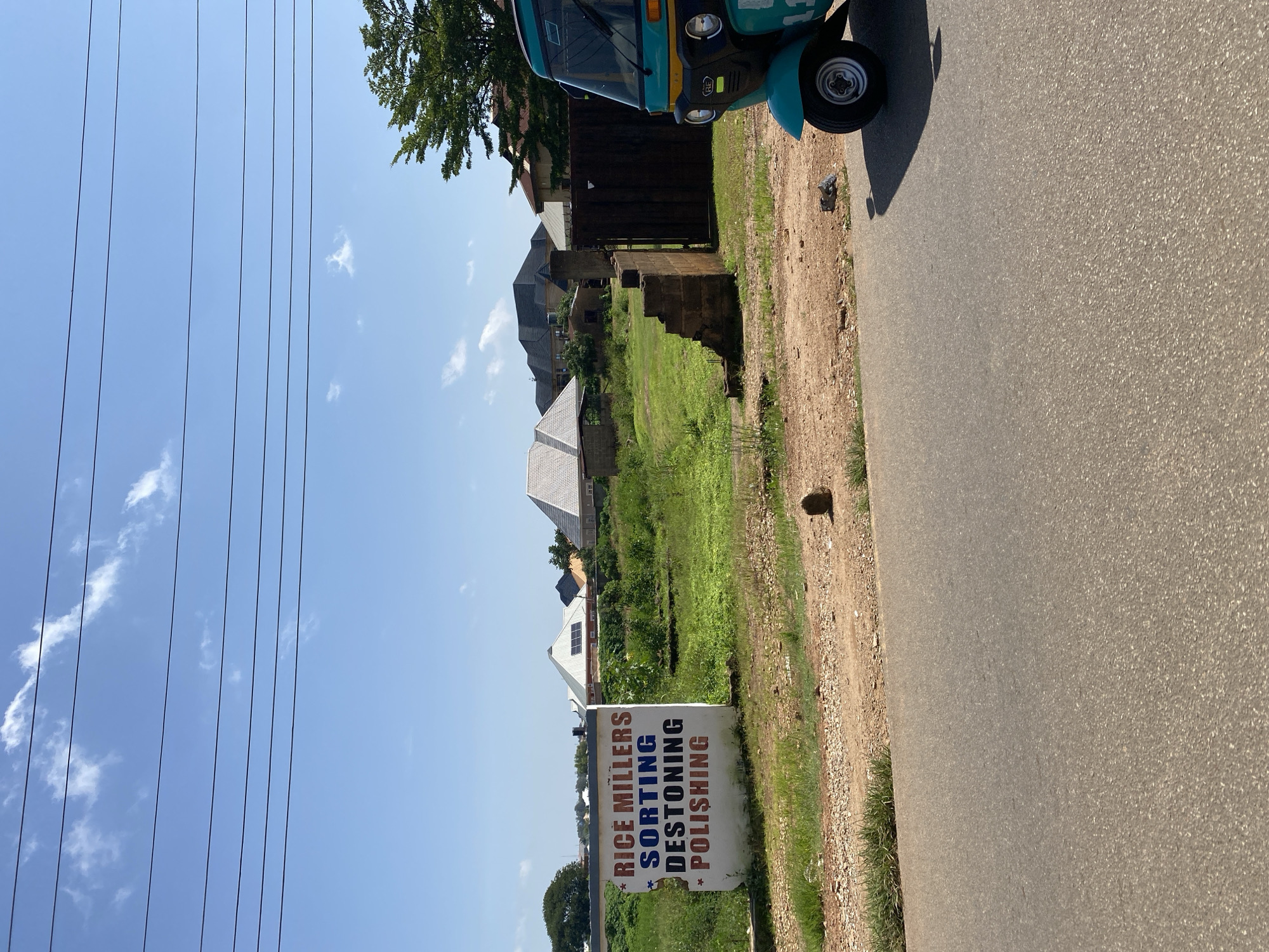 The image depicts a scene in Gura Top, Nigeria, on a clear day with a blue sky and scattered clouds. In the foreground, a paved road runs horizontally across the lower portion of the frame. To the right, a portion of a turquoise and yellow auto-rickshaw is visible, parked on a dirt shoulder.
Behind the road, a white sign is prominently displayed, featuring red text that reads "RICE MILLERS" at the top, followed by blue text "SORTING" and then red text "DESTONING" and "POLISHING". The sign is positioned against a backdrop of a grassy, uncultivated field. In the middle ground, several buildings with modern roofing are visible, suggesting a residential or commercial area. A tall, dark metal gate stands to the right of the sign, partially obscuring a concrete structure. To the far right, a lush green tree reaches into the frame. The overall impression is of a semi-urban or peri-urban environment.