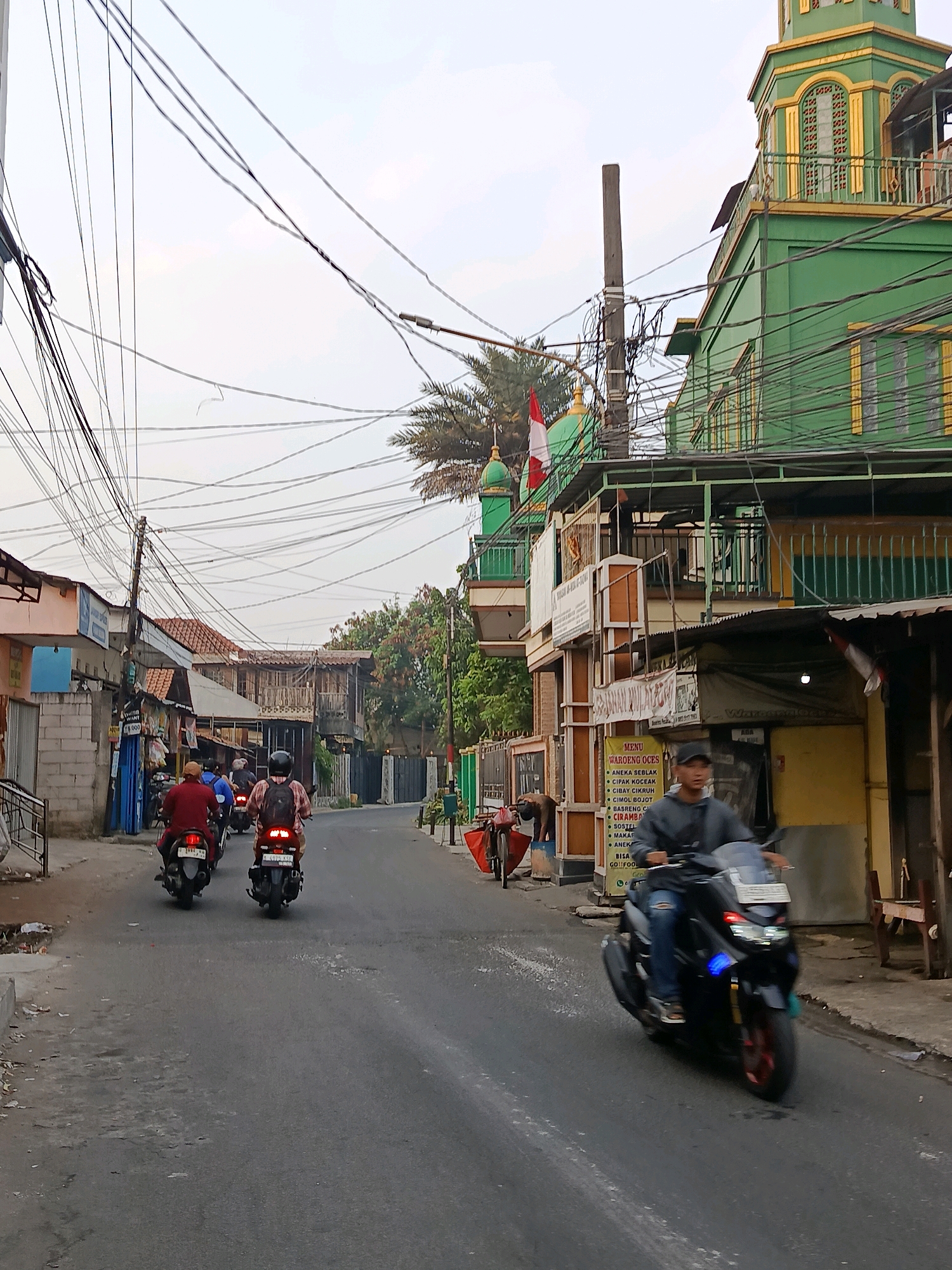 The image shows a street scene in the city of Bekasi, Indonesia. A green building with gold accents, possibly a mosque or community center, is visible on the right side of the frame. In front of this building, there is a yellow storefront with a sign displaying a menu for "WAROENG OCESS". Several people are riding motorcycles on the paved street. Two motorcyclists are in the foreground, with one on a black motorcycle and another on a darker motorcycle. Further down the street, two more motorcyclists are riding away from the camera. The street is lined with buildings and power lines are strung overhead, creating a dense network. Trees and palm trees are also visible in the background. The sky is overcast.