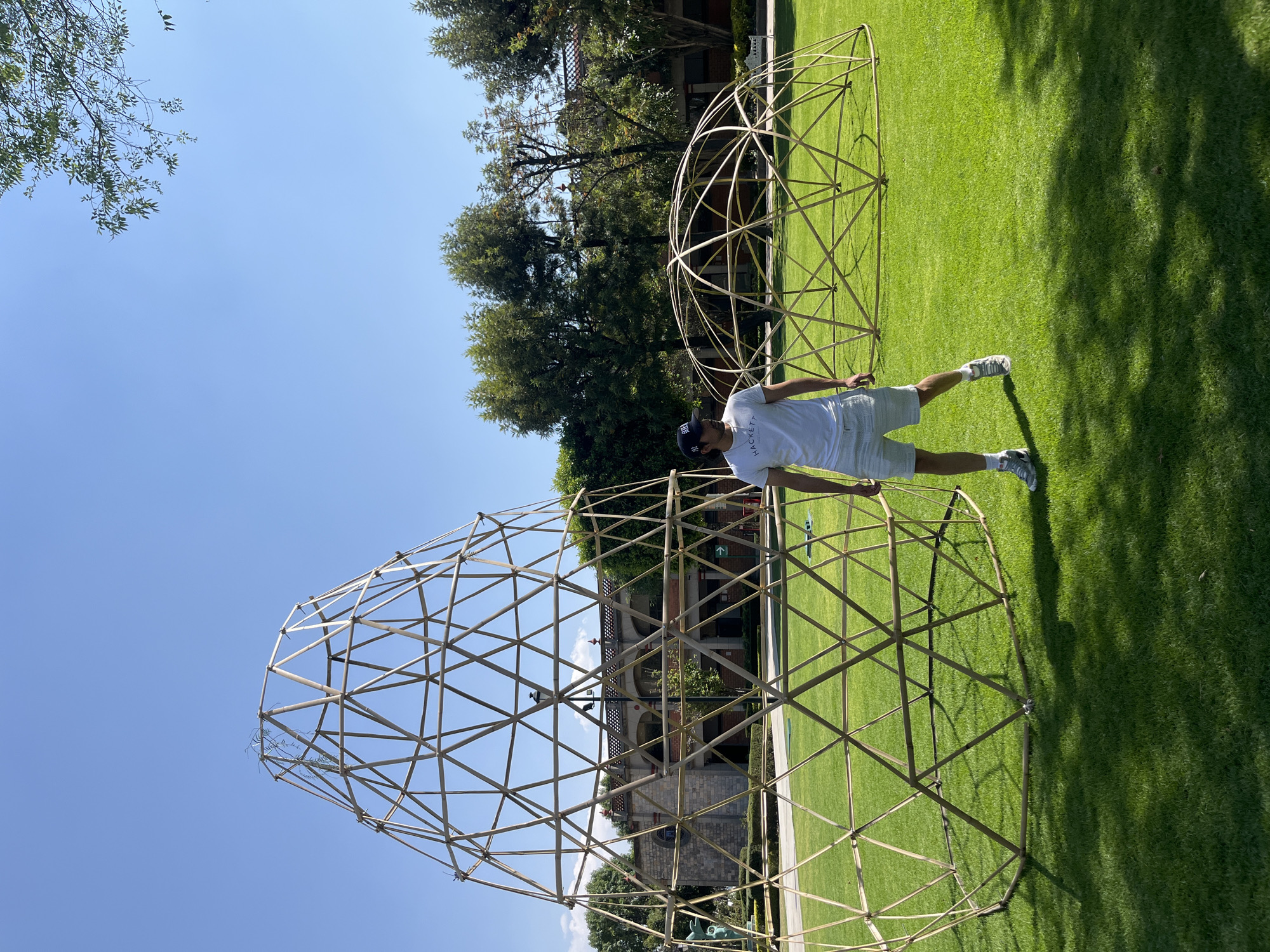 The scene depicts an outdoor area on the campus of the Universidad de las Américas Puebla (UDLAP) in San Andrés Cholula, Mexico, under a clear blue sky. A meticulously maintained green lawn covers the ground, illuminated by direct sunlight, which casts sharp shadows from objects and trees.
Two geometric structures constructed from light-colored poles, likely bamboo, are present on the lawn. The primary structure, positioned on the left, is conical, composed of a geodesic pattern of triangular segments, and stands several meters tall. A smaller, hemispherical geodesic structure, also made of similar materials, is located further to the right and behind the conical structure.
One male individual stands between the two structures. He is wearing a white short-sleeved t-shirt with "HACKETT" printed on the front, light-colored shorts, light grey athletic shoes, and a dark baseball cap. He is oriented towards the right side of the frame, with a neutral stance.
In the background, partially obscured by trees and the structures, is a building with stone walls, a red-tiled roof, and arched windows, consistent with the UDLAP campus architecture. A white directional arrow sign is visible near the base of the conical structure.