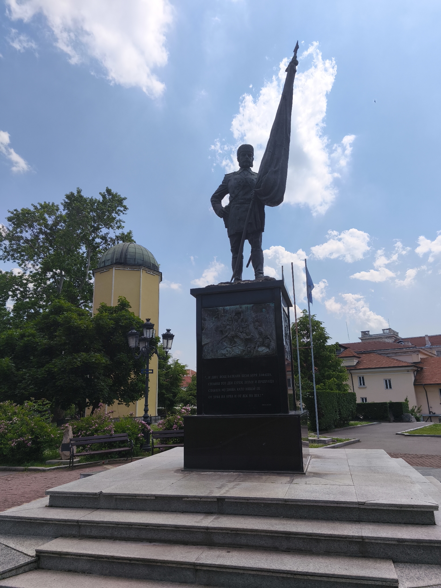 This image captures a wide view of the Monument to the Volunteers (Паметник на опълченците) in Sofia, Bulgaria, specifically located near the Doctor's Garden and the Astronomical Observatory of Sofia University "St. Kliment Ohridski."
The central and dominant feature is a large bronze statue of a male figure in a 19th-century style military uniform, depicted holding a heavy, draped flag pole with his right hand, while his left hand rests on his hip. The figure sports a beard and a military cap. The statue is elevated on a tall, rectangular pedestal constructed from dark, polished stone, possibly granite. This pedestal features intricate bronze bas-reliefs on at least two visible sides, illustrating dynamic battle or historical scenes involving multiple figures. Cyrillic script is etched into the stone on one visible panel of the pedestal.
The pedestal rests on a multi-tiered base of lighter grey stone steps, creating a raised platform from the surrounding ground level. The immediate ground around the monument's base consists of a reddish-brown brick-paved area and lighter grey paved pathways.
In the background to the left of the statue, a distinctive pale yellow building with a dark, spherical domed roof, which is the Sofia University Astronomical Observatory, is visible through and above dense green trees. In front of the observatory and trees, a black multi-lantern streetlamp stands, and a dark wooden bench is partially obscured by lush green shrubs with small pink blossoms.
To the right of the statue, behind a well-maintained, trimmed green hedge, are several buildings with terracotta tiled roofs and light-colored facades, suggesting residential or institutional architecture. Two flagpoles are positioned to the right of the statue's base; one prominently displays a light blue flag, possibly the European Union flag, while the other appears bare or less distinct. More green trees are visible in the distance beyond these buildings.
The scene is illuminated by bright daylight under a clear blue sky, dotted with scattered white cumulus clouds. A small, dark bird is visible in the distant sky above the flag held by the statue.