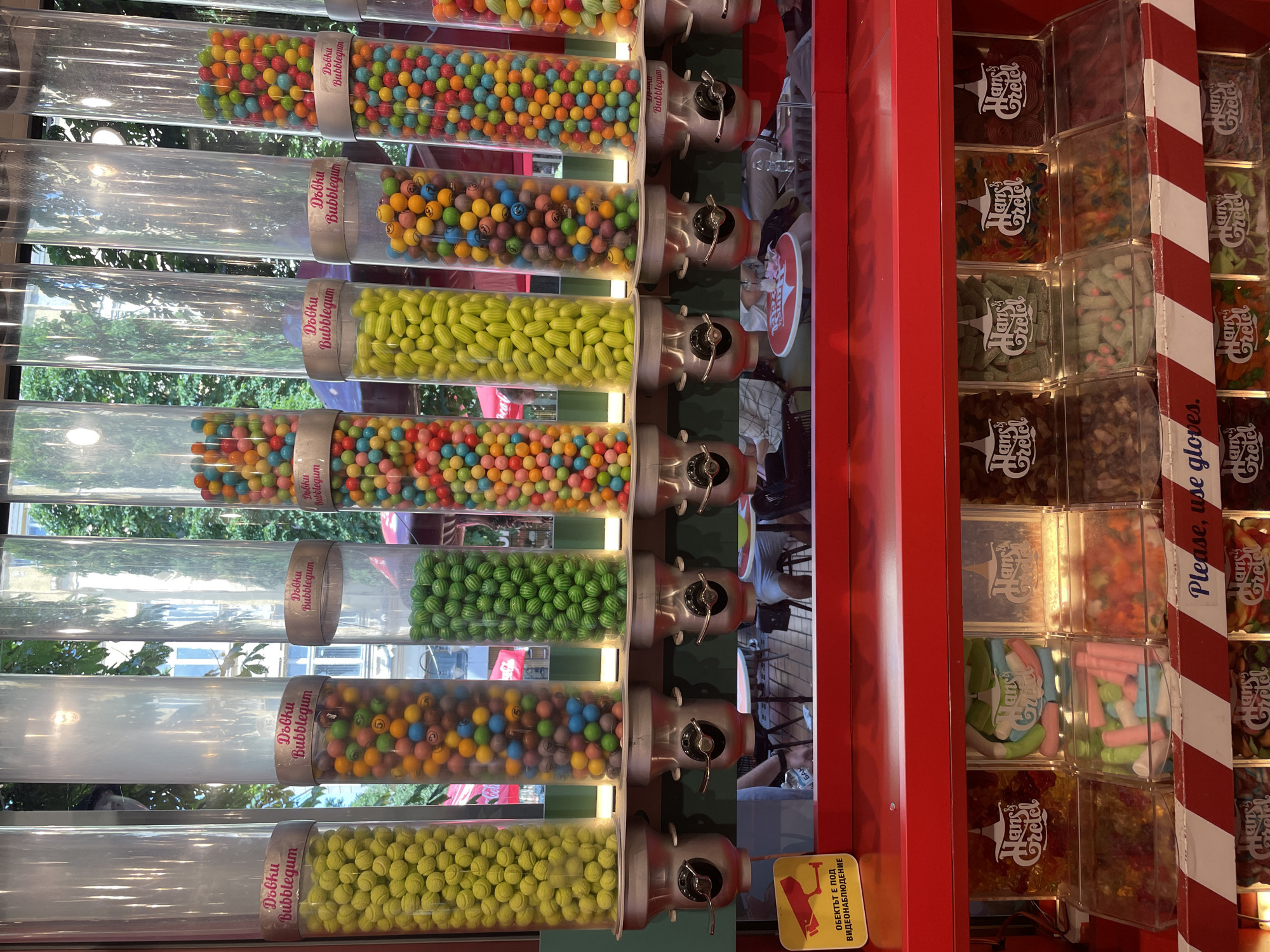 The image displays a candy store setup, specifically a multi-tiered bulk candy dispenser system, located in a commercial area of Sofia, Bulgaria. The upper section features eight transparent cylindrical tubes, each filled with a different variety of gumballs or chewable candies, labeled with pink banners that read "Dobku Bubblegum." From left to right, the visible tubes contain: yellow spherical candies textured like tennis balls; multi-colored smooth spherical candies; green oblong candies textured like watermelons; multi-colored spherical candies, many bearing printed smiley faces; yellow oblong candies; multi-colored spherical candies, many with smiley faces; and two tubes of various multi-colored spherical candies. Each tube is fitted with a metal crank mechanism at its base for dispensing.
Below this, a red counter holds a lower display unit with multiple transparent bins containing various bulk candies, each section branded with the "Hans & Gretel" logo. Visible candies include gummy bears, multi-colored marshmallows (pink, green, white, blue), and assorted gummy shapes and worms. A red and white striped warning sign across the front reads "Please, use gloves."
On the top red shelf to the left, a yellow sign with a black security camera icon states in Bulgarian, "ОБЕКТЪТ Е ПОД ВИДЕОНАБЛЮДЕНИЕ," which translates to "OBJECT IS UNDER VIDEO SURVEILLANCE."
The background, visible through the clear tubes and the shop window, shows an outdoor urban environment with green trees, distant buildings, and a partial red "Coca-Cola" logo. Reflections in the glass reveal parts of an outdoor seating area with tables and chairs, and partially visible human figures seated, suggesting the candy display is situated near an outdoor cafe or restaurant.