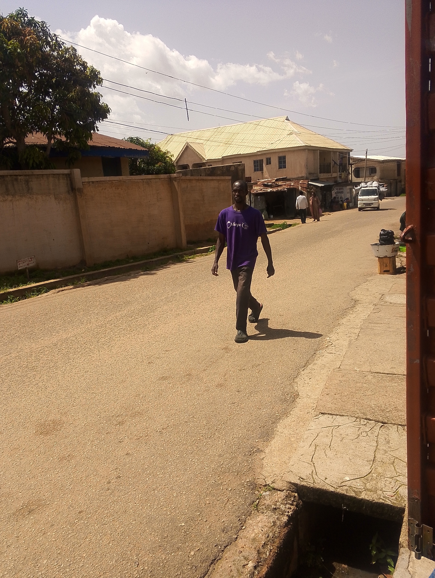 The image depicts a street scene in Jos, Nigeria. A man wearing a purple t-shirt and dark pants walks down the center of a paved road. He is in mid-stride, casting a shadow on the ground. In the background, there are several buildings, some with corrugated metal roofs and others with lighter-colored roofing. A white van is parked on the right side of the road. A concrete wall runs along the left side of the street, with some vegetation growing along its base. The sky is partly cloudy. There is a drain on the right side of the road near the foreground.