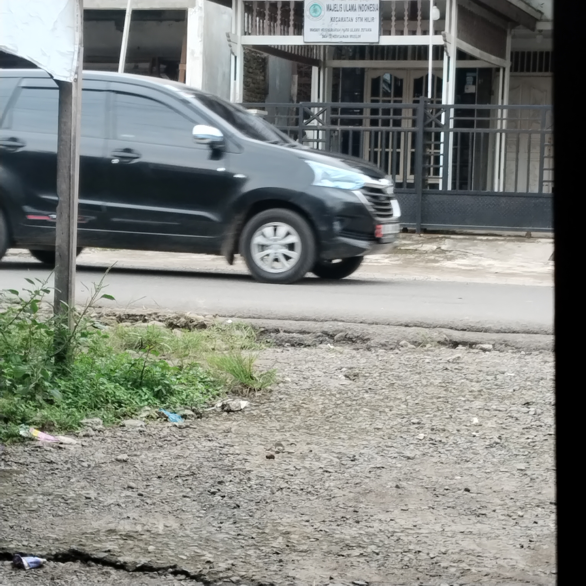 The image shows a black car driving on a road. In the background, there is a building with a sign that reads "MAJELIS ULAMA INDONESIA KECAMATAN STM HILIR." The scene appears to be in a semi-urban or suburban area, with some vegetation visible along the roadside. The road has some cracks and is somewhat uneven. There is also some debris on the ground near the vegetation. The lighting suggests it is daytime.