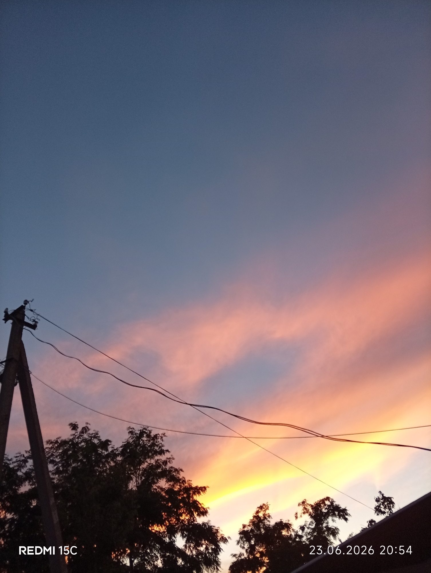 This is a vertical, outdoor, mid-range shot of a sky at dusk. The sky is a gradient of colors, transitioning from a deep blue at the top to soft pinks and oranges nearer the horizon. The colors suggest it is either sunrise or sunset. Silhouetted against the sky are the dark shapes of trees and a wooden utility pole with wires. The utility pole is on the left side of the frame and appears to be made of rough, weathered wood. Several electrical wires stretch across the frame from the pole, disappearing out of view. The trees are dense and leafy, also silhouetted. The overall mood is peaceful and serene, evoking the quiet transition between day and night. Visible text in the bottom left corner reads "REDMI 15C," and in the bottom right corner, the date and time "23.06.2026 20:54" are displayed. The location is Mykolaivka, Ukraine. The weather appears clear, with no visible clouds other than the wisps catching the colorful light.