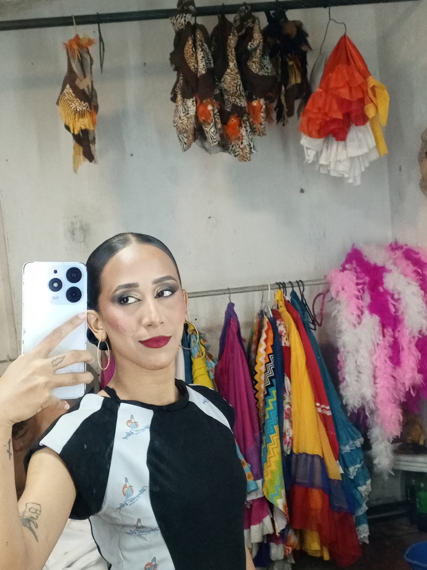 A woman is taking a selfie in what appears to be a dressing room. She has dark hair pulled back, prominent cheekbones, and is wearing dark lipstick and heavy eye makeup. She is wearing gold hoop earrings and a black and white t-shirt. The camera is a white smartphone with multiple lenses on the back. The woman has tattoos on her hand and arm.
Behind her, a rack of colorful clothing is visible. There are several layered, ruffled skirts in various colors, including red, yellow, blue, and pink. There is also a pink and white feather boa. Hanging above the clothing rack are several items with patterned fabrics and fringed edges, possibly headpieces or shawls. An orange and white ruffled skirt is visible. The wall is white and appears to be a simple, undecorated surface.
The location is implied to be Porlamar, Venezuela.