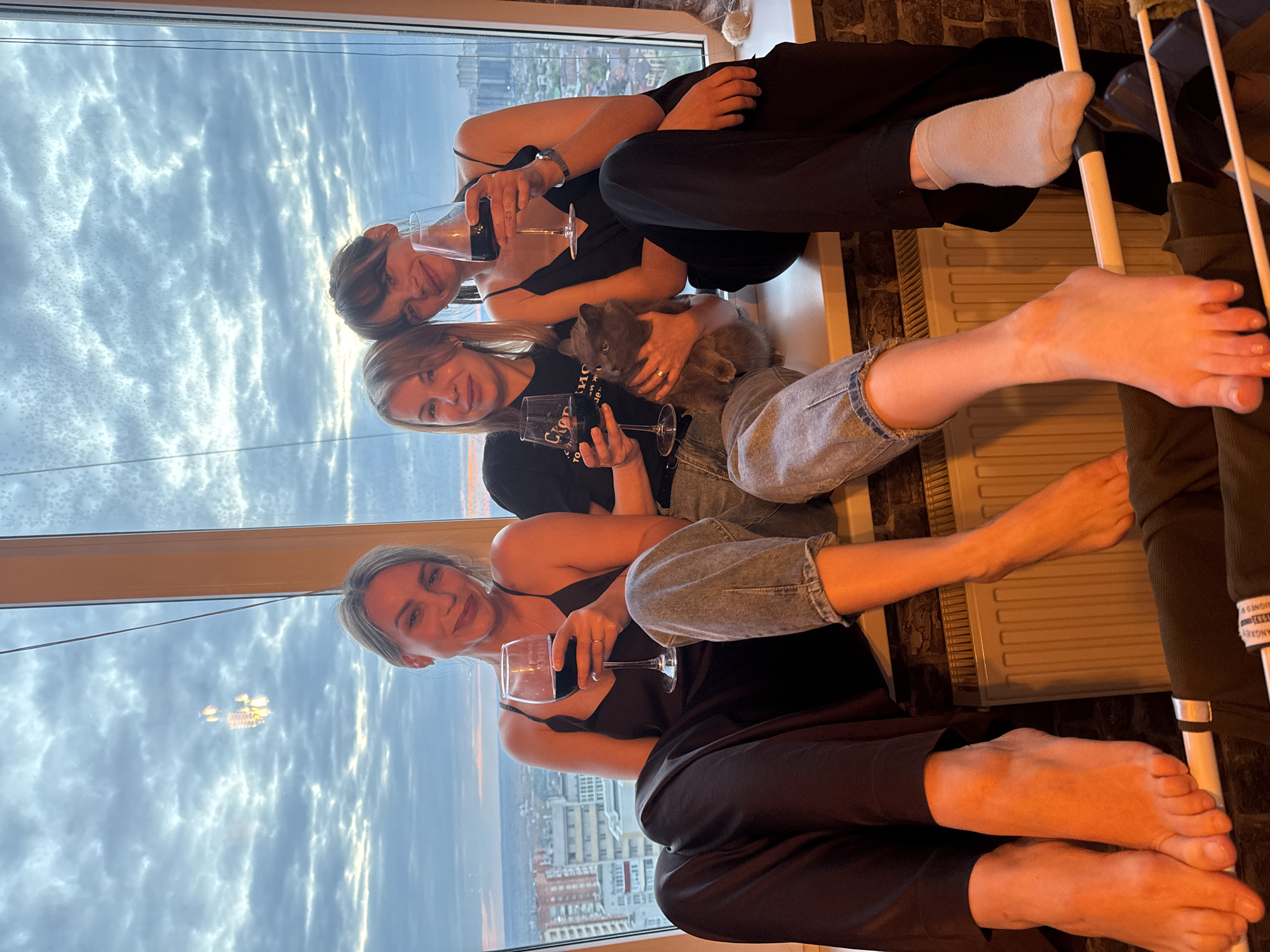 Three women are sitting by a window overlooking Rostov-on-Don, Russia, drinking wine. The woman at the top is wearing a black dress and white socks. The woman in the middle has blonde hair and is holding a grey cat. The woman at the bottom is wearing a black tank top and black pants.