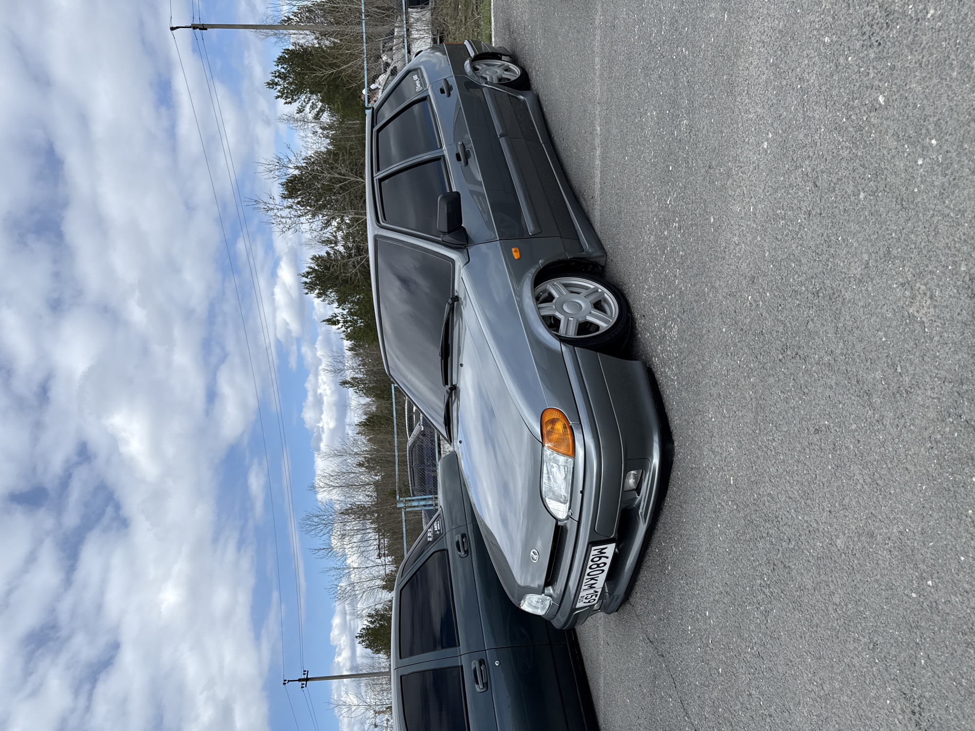 A gray Lada sedan is parked on a paved surface. The license plate reads "M680KM 59," which indicates the vehicle is registered in the Perm Krai region of Russia. A darker vehicle is partially visible next to the Lada. A wooded area and a blue sky with clouds are in the background. Based on the license plate, the location is within Perm Krai, Russia, and likely in or near the city of Perm.