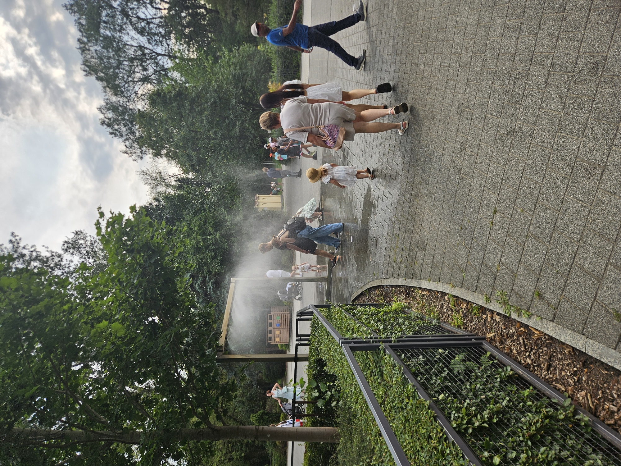 The image depicts an outdoor scene in a park-like setting with a misting station in operation. Several people are visible, some interacting with the mist, while others walk past. A paved pathway curves through the scene, bordered by greenery and a metal fence. The sky is overcast with clouds. Based on the architectural elements of the misting station and the surrounding vegetation, this scene is located at the Zoological Garden in Warsaw, Poland.