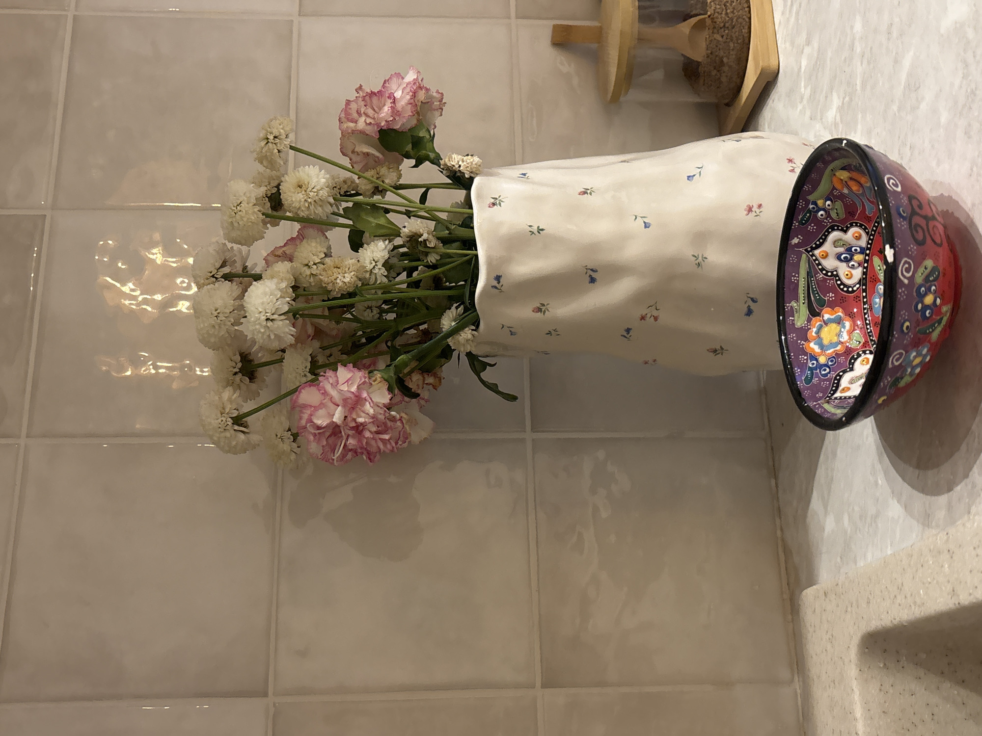A vase of flowers sits on a tiled surface next to a bowl. The vase is off-white with a small floral pattern. The flowers in the vase are a mix of white and pink carnations and other white flowers with small heads. The bowl is decorative with intricate colorful patterns. To the left of the vase is a wall covered in square, cream-colored tiles. The environment suggests a bathroom or kitchen countertop, and the details suggest a domestic interior. There is no information in the image to suggest this scene is located in Krasnoyarsk, Russia.