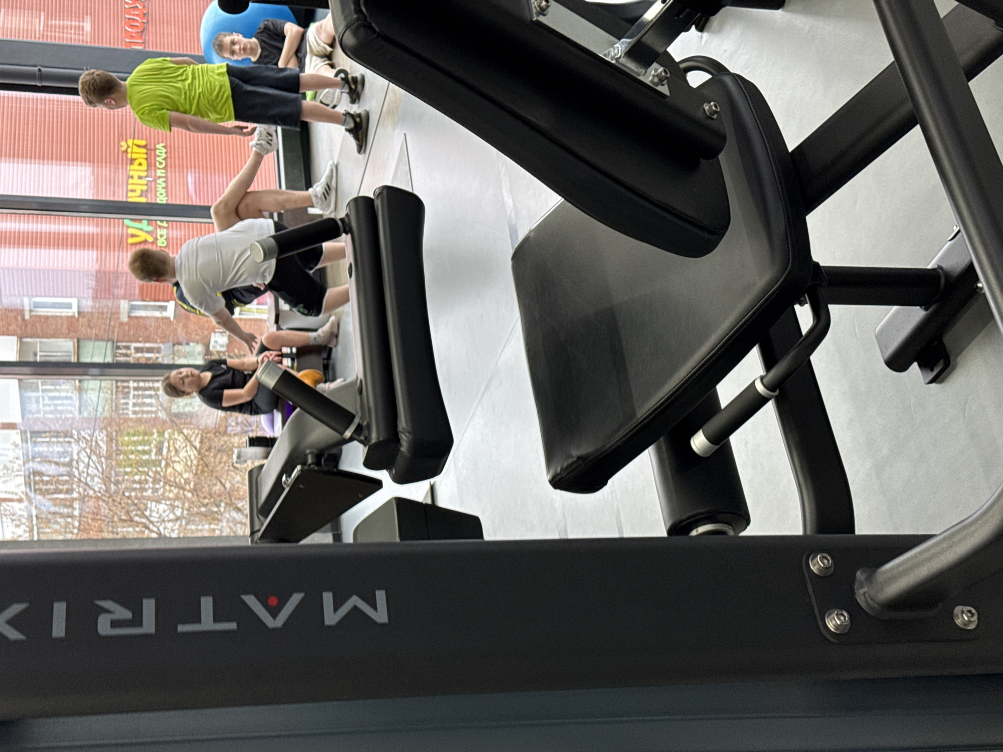 A gym scene shows a Matrix exercise machine in the foreground. Through the gym windows, a view reveals buildings, trees lacking leaves, and signage with Cyrillic script, likely in Votkinsk, Russia. Several individuals are in the background performing exercises. One person lies on a blue exercise ball, while others stretch or use other equipment. The lighting suggests it's daytime.