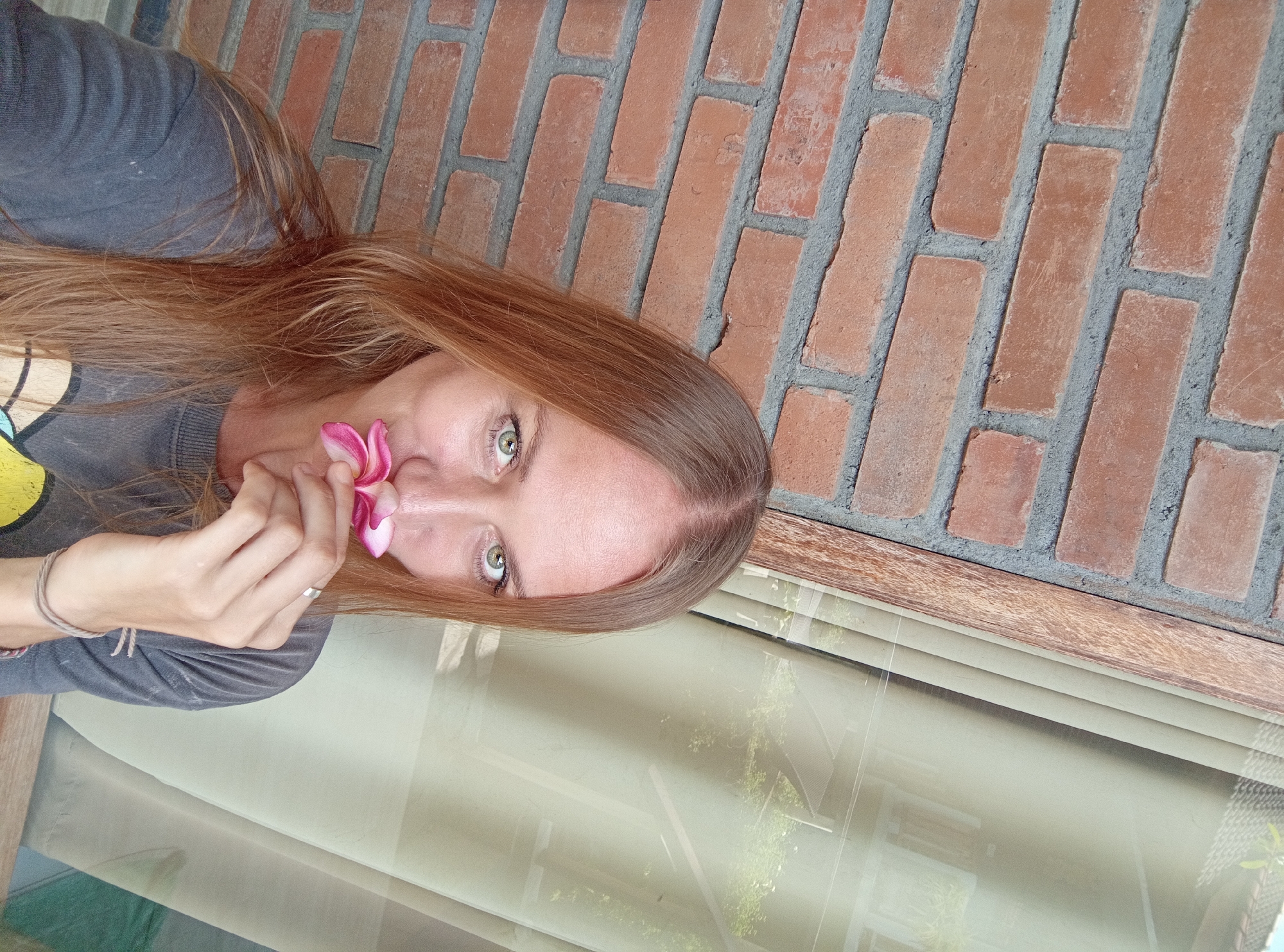 The image depicts a woman with light brown hair and green eyes, holding a pink plumeria flower near her face. She wears a gray sweatshirt and a ring on her finger. The background features a red brick wall and a window reflecting an exterior scene with greenery. The location is North Kuta, Indonesia.