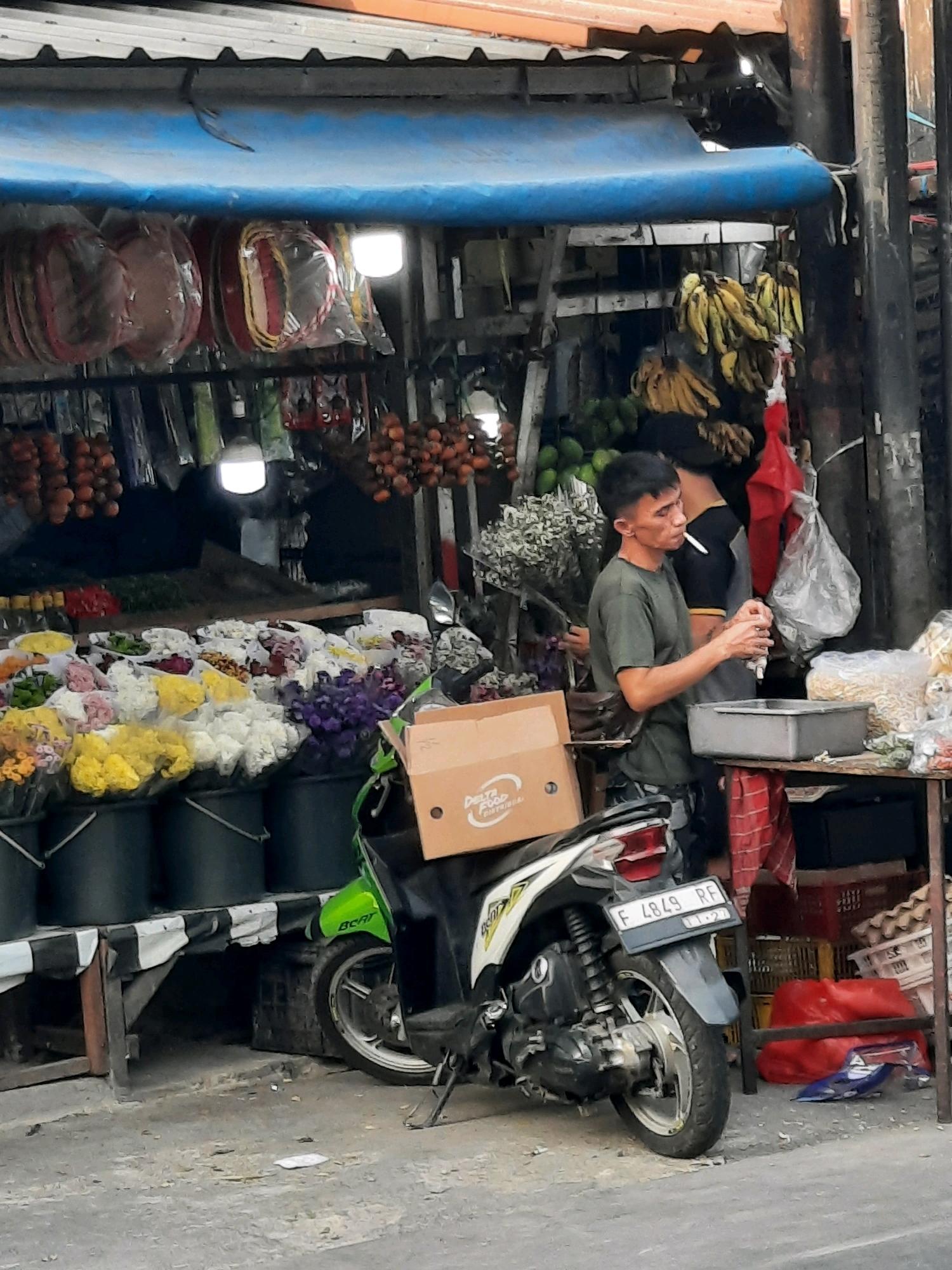 The image depicts a street market scene in East Jakarta, Indonesia. A man wearing a green t-shirt is standing and smoking a cigarette, holding a small white object in his right hand. He is positioned near a grey rectangular container and a table with various food items. Behind him, another person in a black shirt is partially visible. In the foreground, a green and white motorcycle is parked. The market stall features an assortment of items, including bananas hanging from the stall, various flowers in buckets, and produce. The stall is covered by a blue tarp. The ground is paved, and there is debris scattered around. The overall impression is a bustling, typical street market environment.