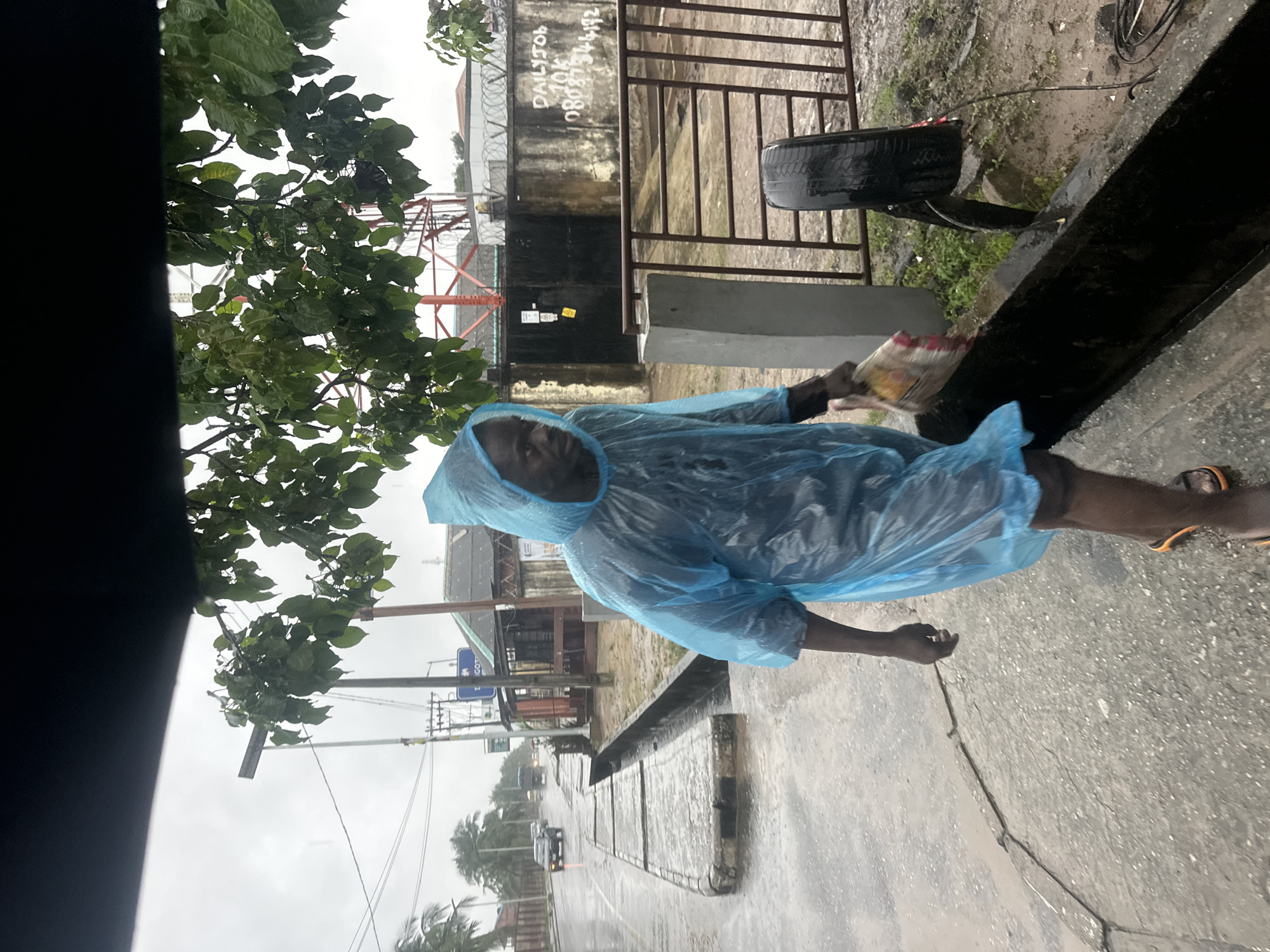 A Black man wearing a light blue disposable poncho walks on a wet concrete sidewalk in what appears to be the city of Badagry, Nigeria. The man is carrying a multi-colored bag in his right hand. He is wearing dark-colored shorts and orange and black sandals. To his left, a street is visible with cars driving in the distance, and it appears to be raining. A concrete drainage ditch runs alongside the sidewalk. Behind the man, a metal fence with barbed wire is visible, with a wall and buildings beyond. There are trees with green leaves hanging over the scene from the top right. A tire is leaning against the fence in the background. The sky is overcast and gray. The ground is wet, indicating recent or ongoing rain.