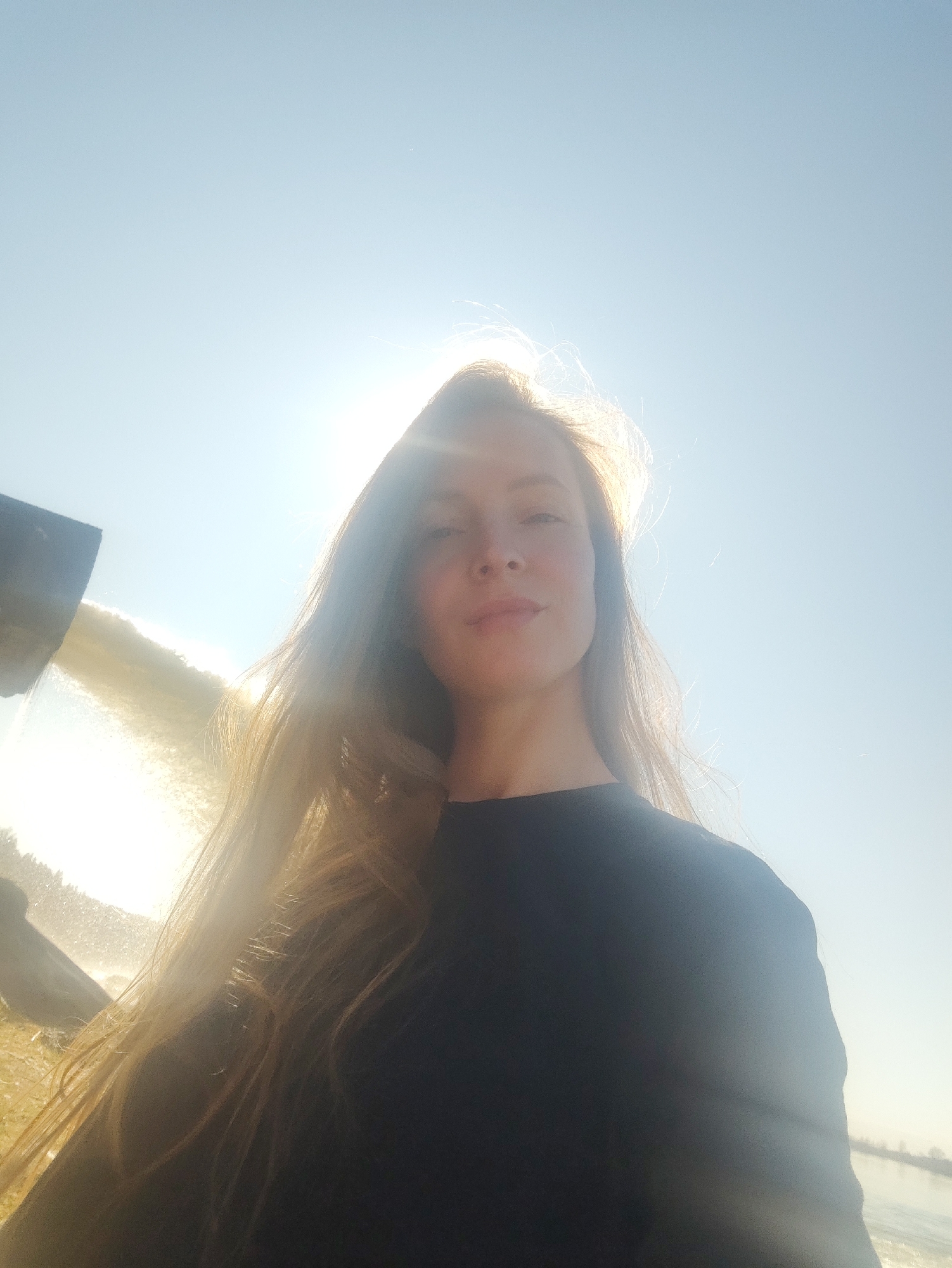 A young woman with long, light brown hair is posing for a selfie-style photograph outside. She is wearing a dark-colored top. The background is a bright, sunny sky with a body of water and what appears to be a small waterfall to the left of the woman. The location is Chernushka, Russia.