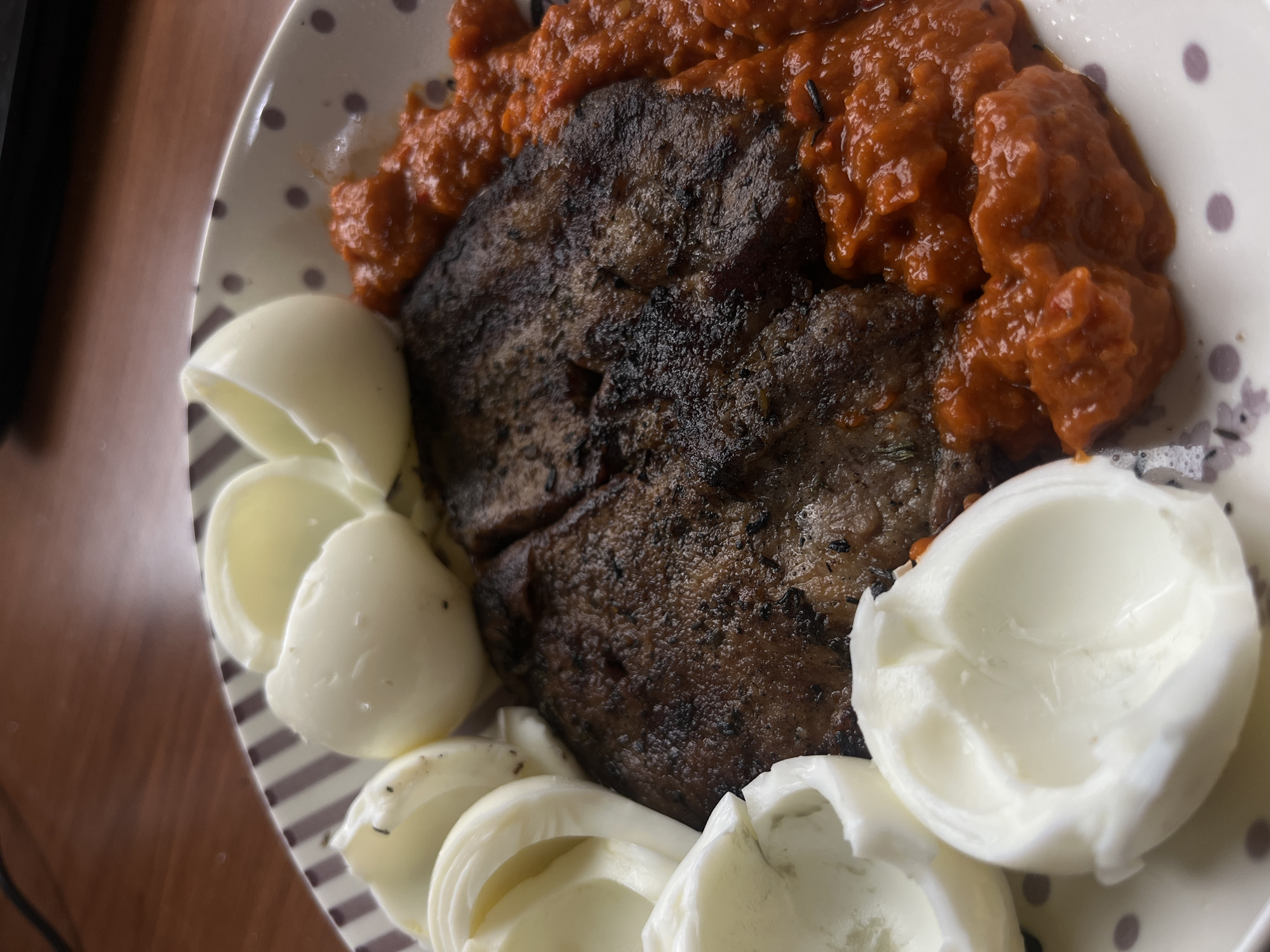 A close-up shot displays a plate filled with food. There are two pieces of what appears to be cooked meat, possibly liver or steak, covered with a generous portion of a thick, reddish-orange sauce. Surrounding the meat are several halved, hard-boiled eggs. The plate itself has a white base with a pattern of brown polka dots and floral designs. The setting lacks explicit indicators of location, and there is no clear evidence to suggest a specific city such as Plovdiv, Bulgaria.
