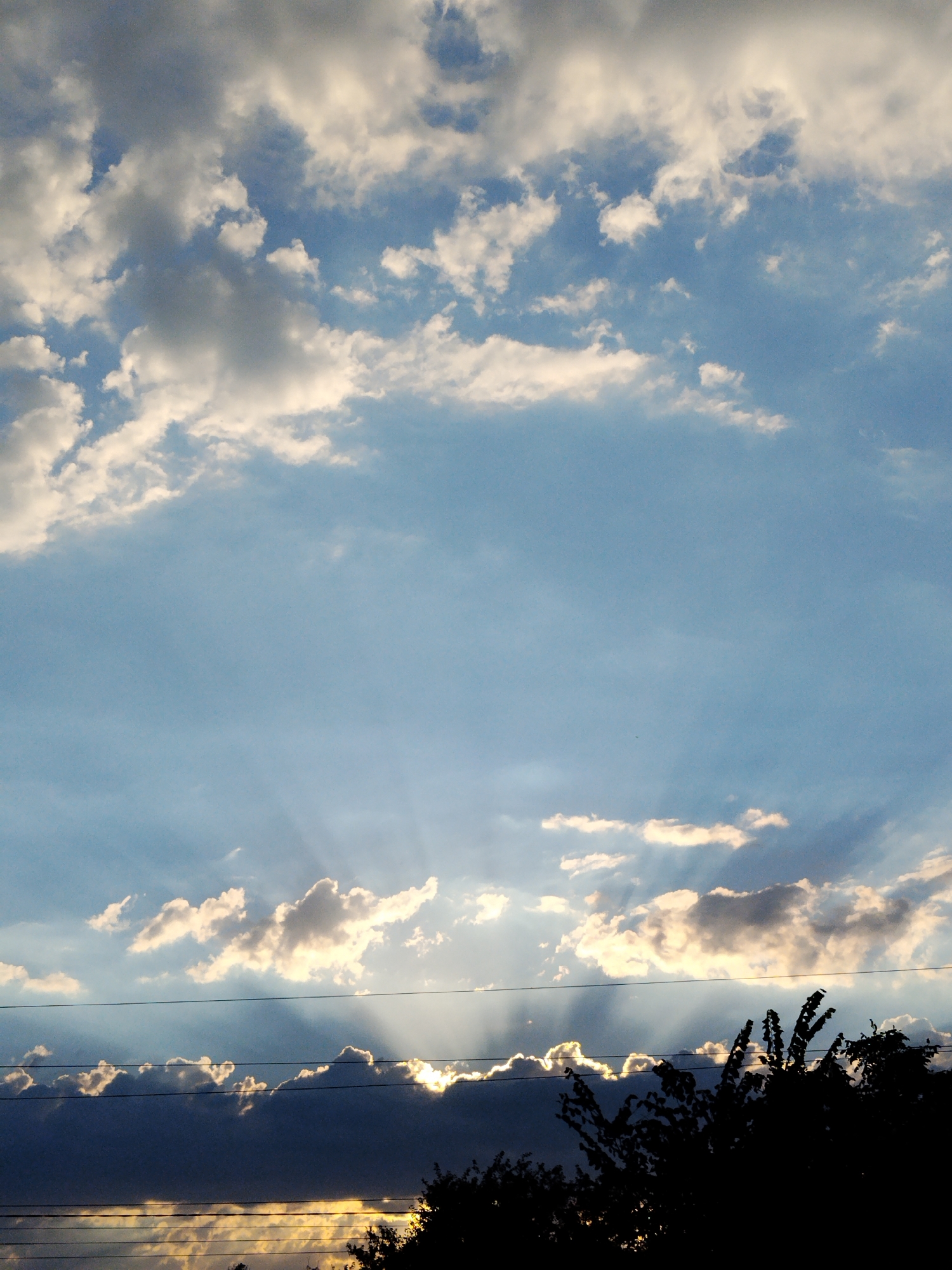 A photo taken in Ustyvytsia, Ukraine, depicts a sky filled with clouds and crepuscular rays. The sun is setting, casting rays of light through breaks in the clouds. The sky is a clear blue, with white and gray clouds scattered across it. In the foreground, there are silhouetted trees and power lines. The overall scene is one of natural beauty and tranquility.