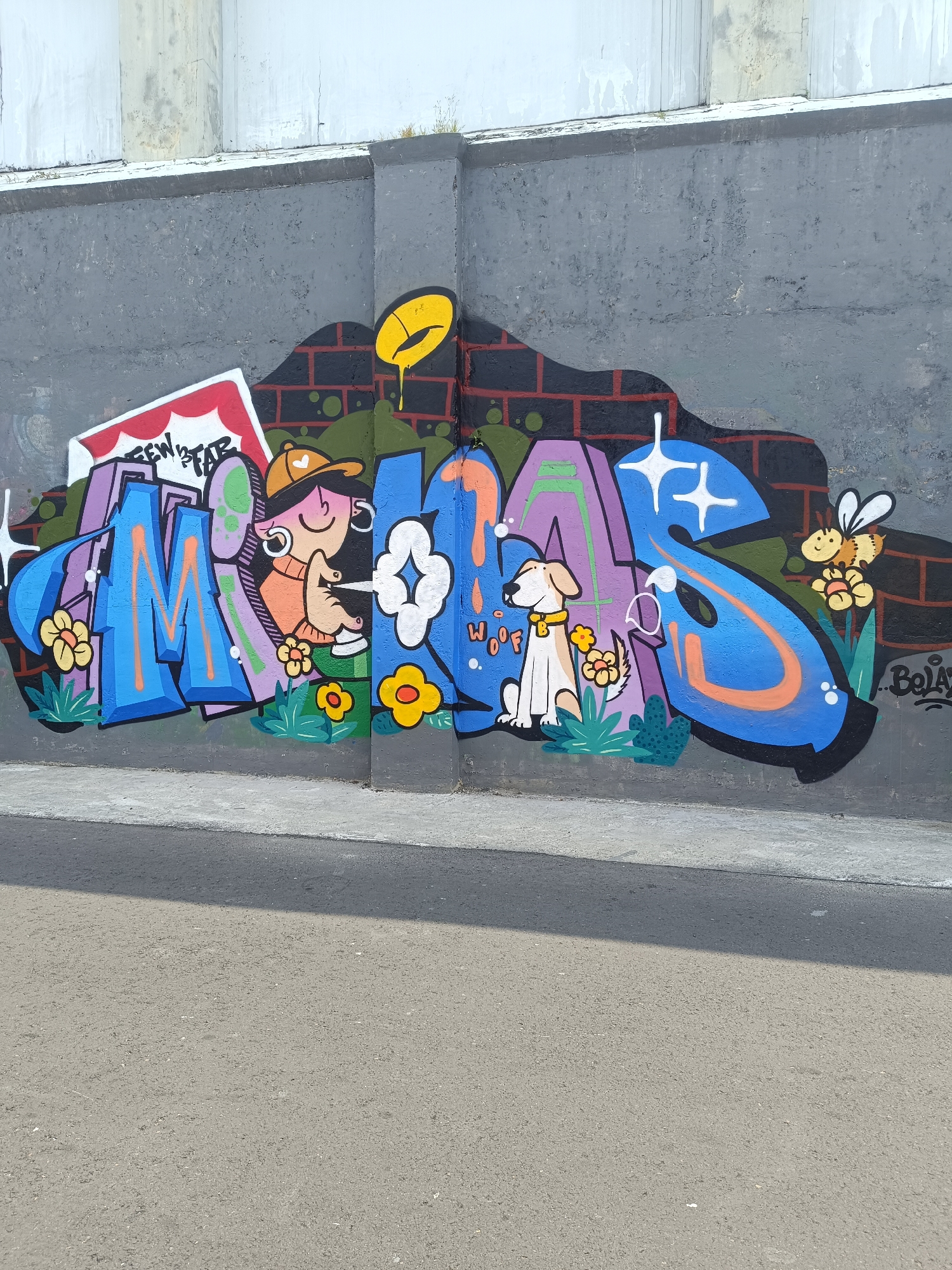 The image depicts a section of a graffiti mural on a grey concrete wall in Yogyakarta, Indonesia. The mural features large, colorful lettering that appears to spell "MAFS." The letters are filled with vibrant blues, purples, and oranges, outlined in black. The mural incorporates various cartoonish elements: a figure with an orange cap and earrings, a white dog with a yellow collar, and a bee with orange and yellow stripes. The dog is positioned next to the letter "S," and the word "WOOF" is written in small, orange letters near it. The bee is located to the right of the "S," hovering near some yellow flowers. The background of the mural includes a brick pattern and green foliage. There is also a yellow oval shape with a drip design at the top of the mural, to the left of the letters. The wall itself shows signs of age and weathering. In the foreground, a grey asphalt road runs parallel to the wall, with a concrete sidewalk or elevated surface above it. The lighting suggests it is daytime, with a shadow cast on the road from an unseen object.