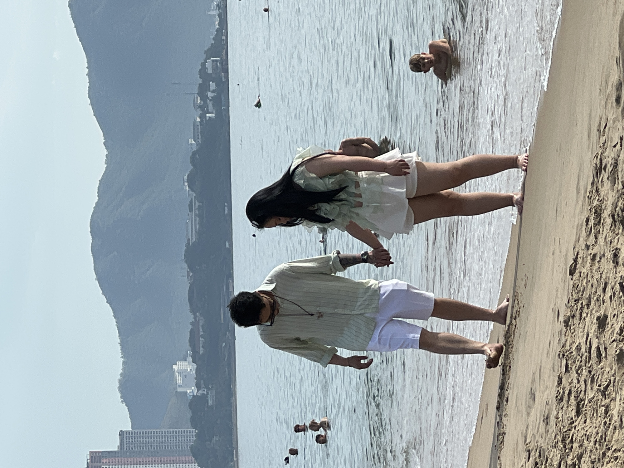 Two individuals, a man and a woman, are walking hand-in-hand on a sandy beach in Nha Trang, Vietnam, with the ocean before them and a cityscape and mountains in the background. The man, on the left, wears a light green striped long-sleeved shirt and white shorts, with sunglasses on his head and a necklace. He has tattoos on his left arm and is looking down. The woman, on the right, is wearing a light green ruffled top and white shorts, with her dark hair long and flowing. She is looking down and to her left. Several other people are visible in the shallow ocean water, with some swimming and one child in the foreground of the water, looking towards the camera. The water is calm with small waves lapping the shore. In the distance, buildings and tall structures are visible on the coastline, with lush green mountains forming the backdrop. The sky is overcast and hazy.