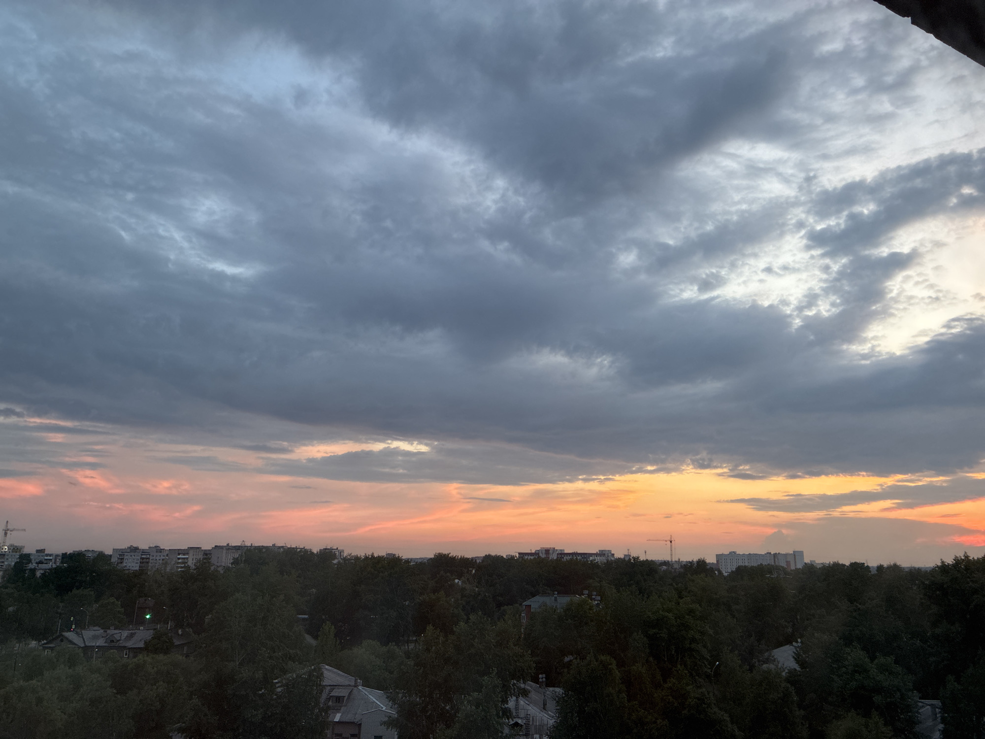 The image shows a wide-angle view of a city skyline at sunset. The sky is filled with dramatic, gray clouds that transition to hues of orange and pink near the horizon. Below the sky, a dense canopy of dark green trees dominates the midground, interspersed with the roofs of buildings. The buildings appear to be a mix of residential blocks and older structures, with some evidence of cranes in the distance, suggesting ongoing construction. The overall impression is of an urban environment during twilight.