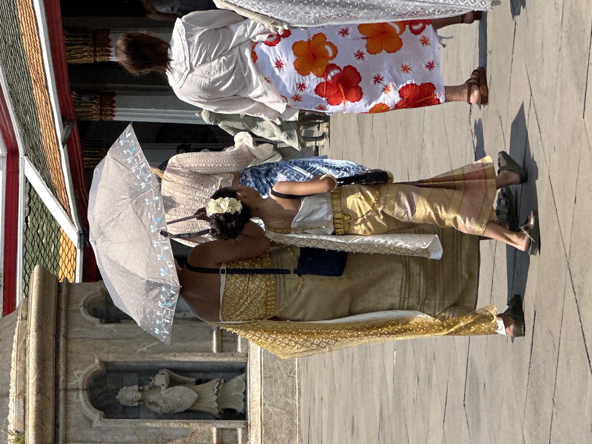 The image shows two women in traditional Thai clothing walking on a paved surface in Bangkok, Thailand. One woman is wearing a long, gold-colored dress with an intricate pattern and a white sash. She is holding a light-colored umbrella with a design of small dots and floral motifs. A small, dark-colored bag is visible at her waist. The other woman is wearing a white top and a skirt with a white background and red and orange floral patterns. Both women are wearing sandals. In the background, there is a building with ornate architectural details, including a red and gold roof and a niche containing a white statue. The paved surface is marked with shadows.