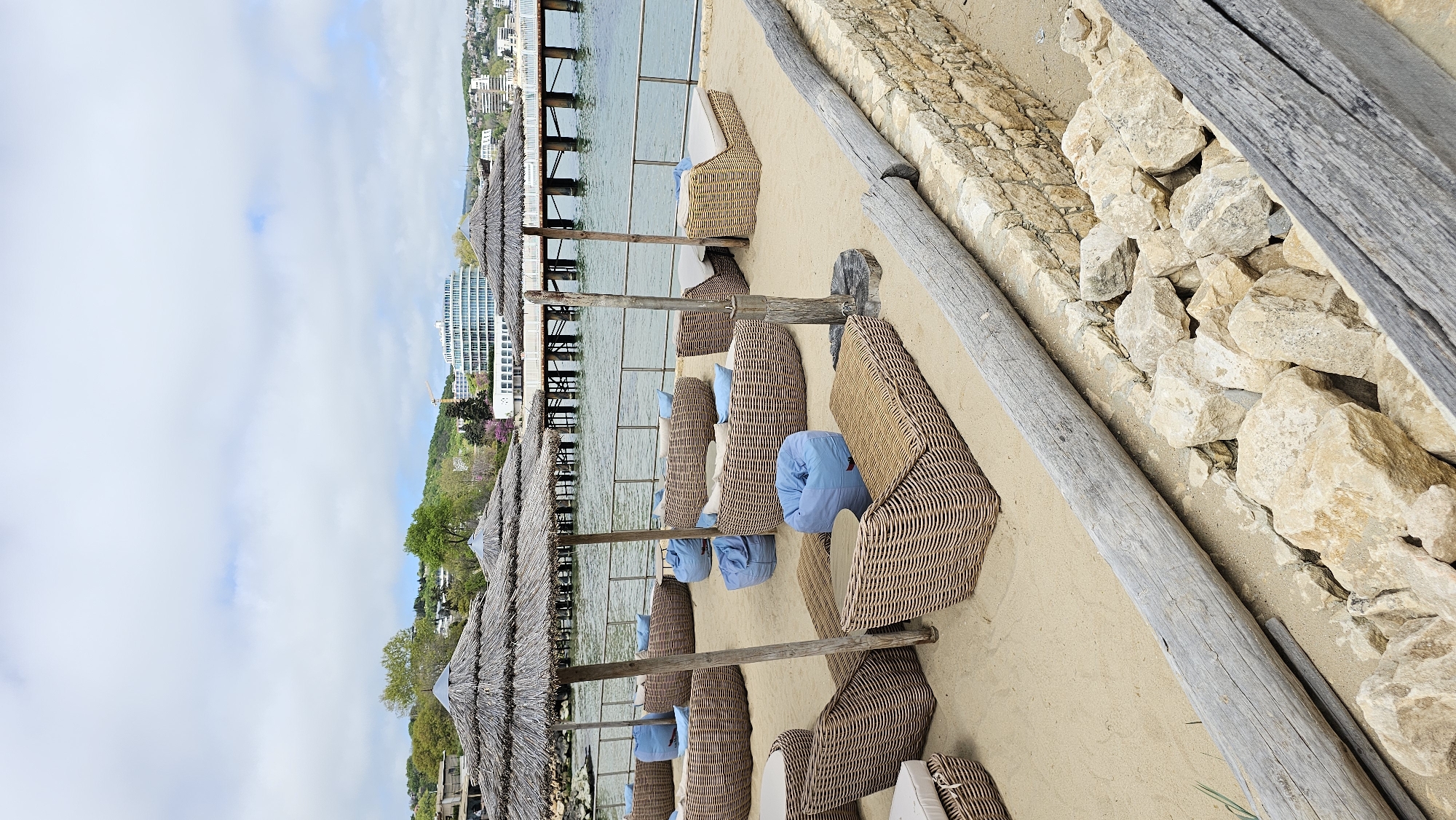 The image depicts a beach scene in Varna, Bulgaria. Several lounge chairs with blue and white cushions are arranged under thatched umbrellas on a sandy beach. In the background, a pier extends into the water, and buildings are visible along the coastline. A stone wall and wooden logs border the beach area. No people are visible in the immediate scene.