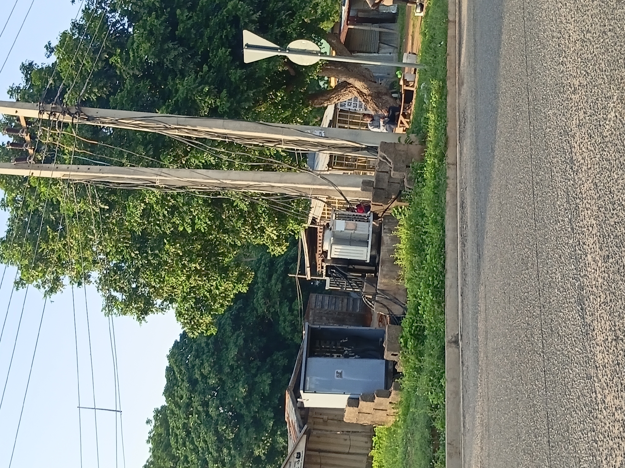The image depicts an outdoor scene in Jalingo, Nigeria, featuring an electrical transformer unit. The transformer is housed in a blue metal enclosure, which is partially open, revealing internal components. The enclosure is situated next to a stack of concrete blocks. In the background, there are various structures, including what appear to be small shops with corrugated metal roofs. Two concrete utility poles with a dense network of electrical wires are prominent in the center of the image. A large tree with thick foliage stands behind the utility poles. Several individuals are present in the scene. One person is seated on a wooden bench near the transformer. Another person is visible near the blue enclosure, and others are further in the background. Green grass and other vegetation surround the transformer and structures. A traffic signpost with a triangular and circular marker is visible to the right of the utility poles. The ground in the foreground is an asphalt road.