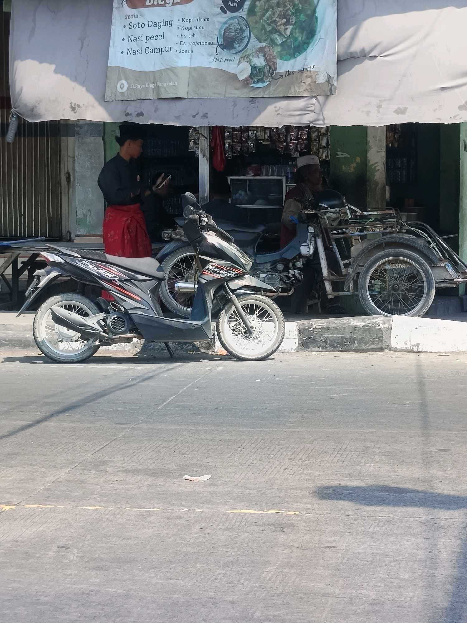 In Blega, Indonesia, a street scene unfolds under bright daylight. A young man wearing a black shirt and a red sarong stands facing a mobile phone, his gaze directed downwards. To his right, an older man, also dressed in darker attire and a white skullcap, is seated on a tricycle-like vehicle. The vehicle, a three-wheeled motorcycle, is parked at the curb. In the foreground, a modern motorcycle, black with red and silver accents, is positioned parallel to the curb, its wheels turned towards the street. The background reveals a storefront with a large banner displaying a menu of food and drinks, including "Soto Daging," "Nasi Pecel," and "Nasi Campur." Products are displayed on shelves behind the counter. The street itself is paved, with a distinct curb separating it from the sidewalk area where the vehicles are parked. The overall atmosphere is that of a typical roadside establishment in a bustling Indonesian town.