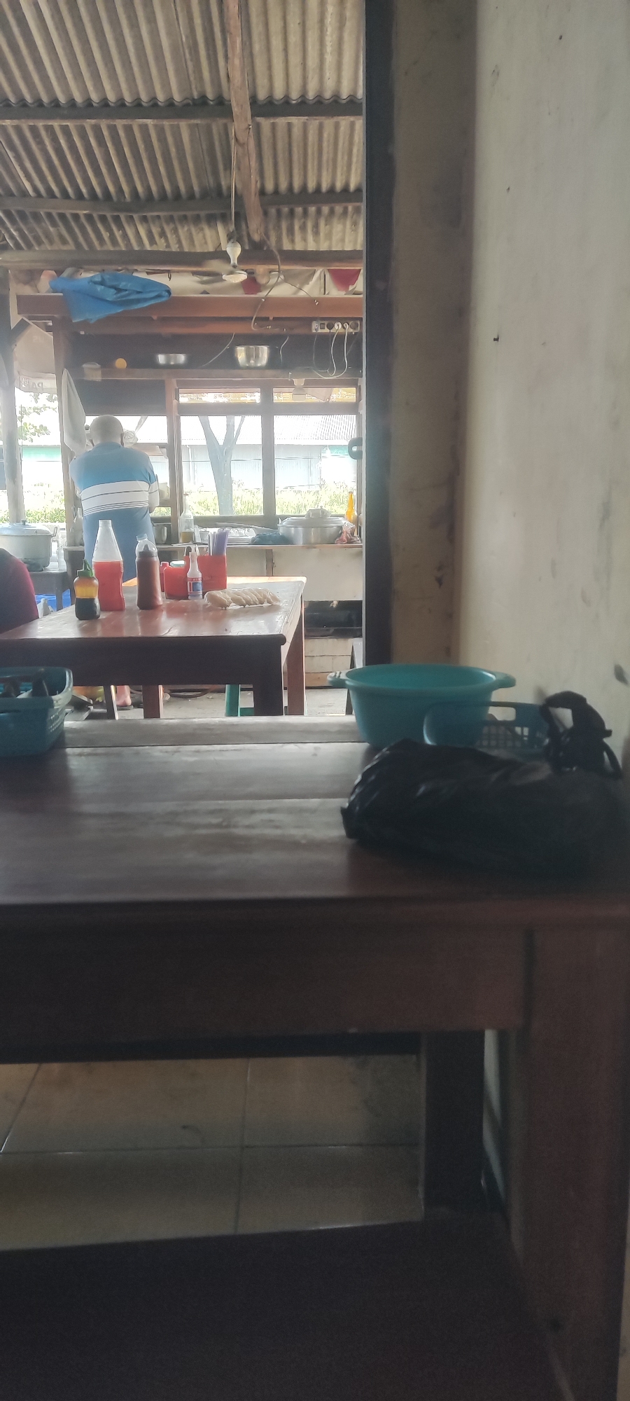 The image depicts the interior of a food stall in Pangenan, Indonesia. A person wearing a blue and white striped shirt is visible in the background, appearing to be working. On a wooden table in the foreground, there are several bottles of condiments, a red container with utensils, and a plate of food. In the right foreground, a large blue plastic basin and a black bag are placed on the table. The stall has a corrugated metal roof, supported by wooden beams. The overall environment suggests a casual, open-air dining or preparation area.