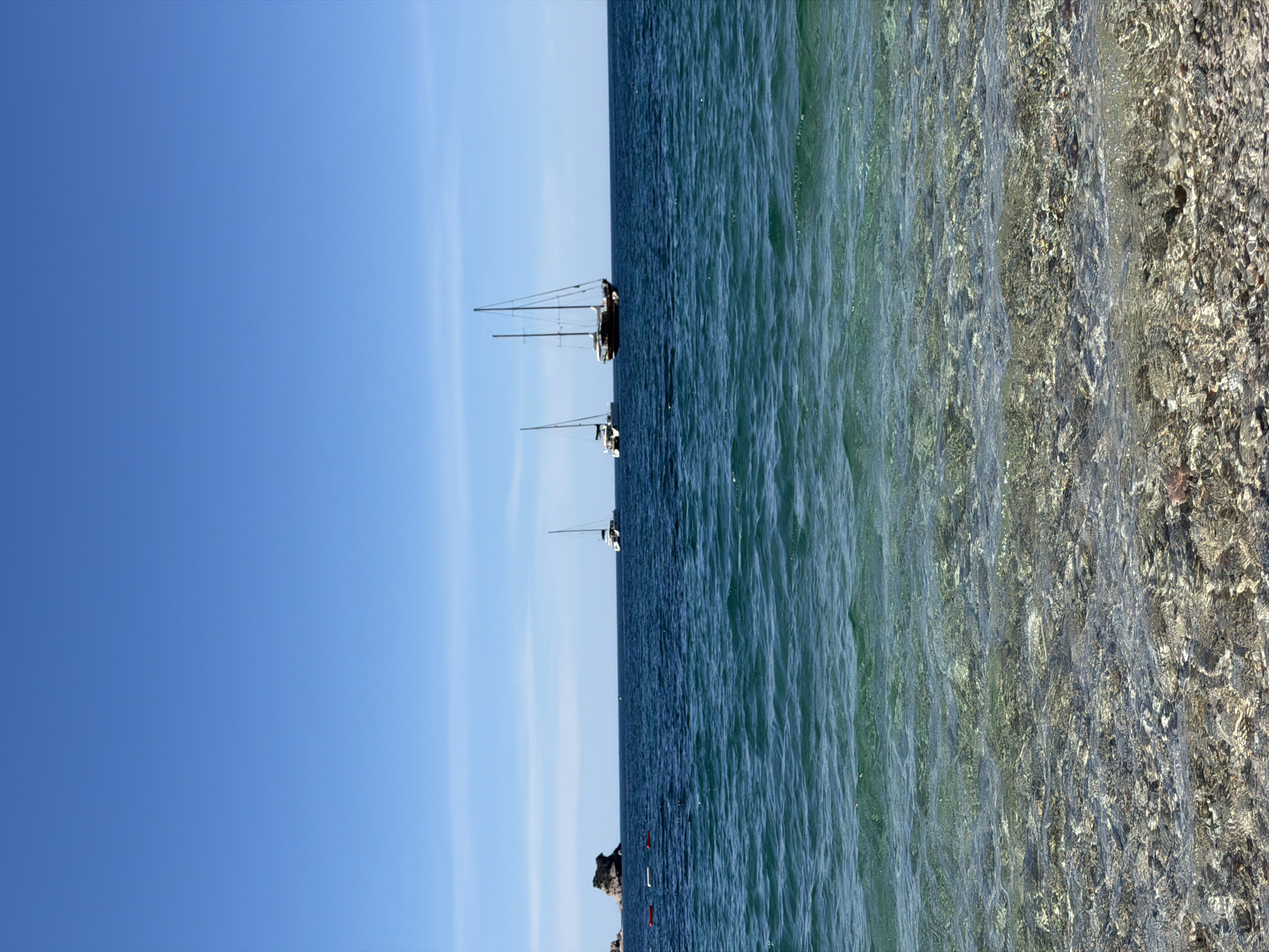The image depicts a scene in Budva, Montenegro. The primary elements are the sky, the sea, and the seabed. The sky is a clear blue with some light clouds. The sea is a mix of darker and lighter blues. There are three sailboats visible on the horizon. The seabed is clearly visible near the shore, with a mixture of sand and pebbles. No people are clearly visible in the image.
