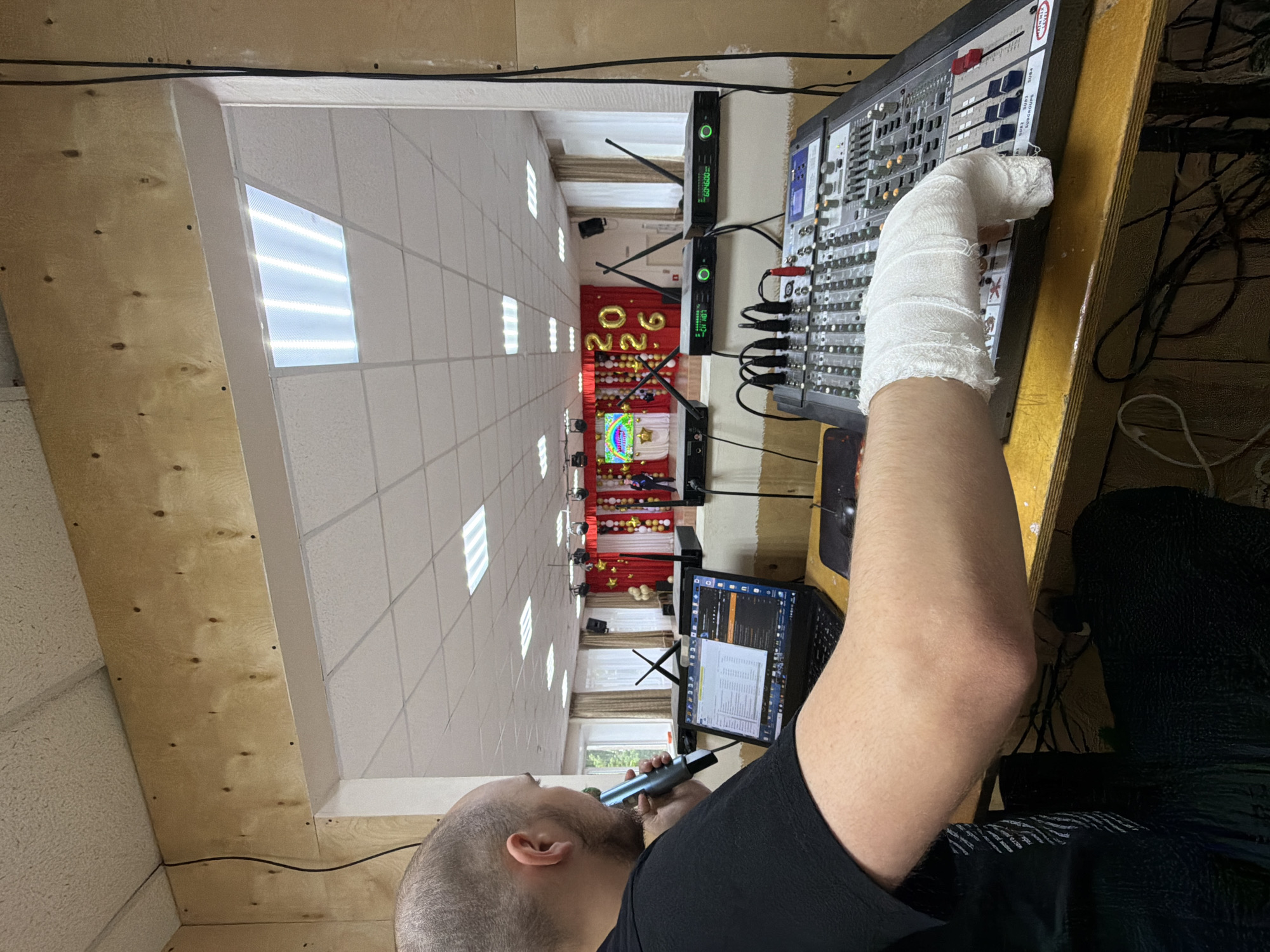 A man stands inside a room in Izhevsk, Russia, facing a sound mixing console. His right arm is in a white cast, and he holds a microphone to his mouth with his left hand. A laptop sits on a table next to the console. The room is decorated with gold balloons spelling "2026."