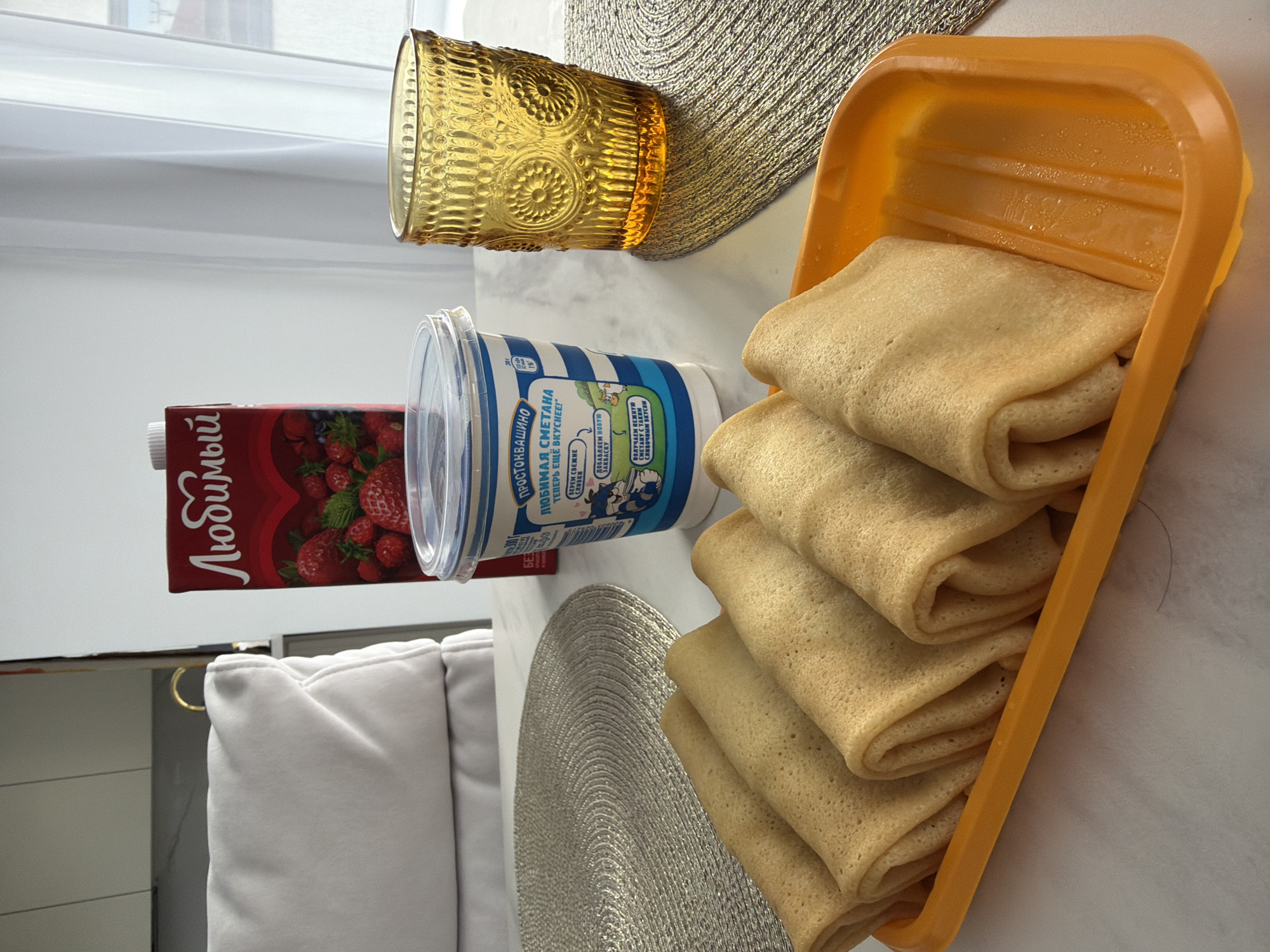 Here is a forensic description of the image:
The image shows a table setting with various food items, likely in a home in Молодёжный, Russia.
**Objects:**
* A stack of folded pancakes in an orange serving dish.
* A container of sour cream labeled "Prostokvashino" (in Russian).
* A carton of strawberry juice labeled "Lyubimy" (in Russian).
* A decorative amber-colored glass.
* Two woven placemats.
* A portion of a white cushioned chair is visible.
**Environment:**
* The setting appears to be indoors, likely on a table near a window, indicated by the presence of sunlight and window frame in the background.
**Summary:**
The image depicts a breakfast or brunch scene with pancakes, sour cream, and strawberry juice. The Russian labels indicate the location is in Russia, possibly in a home.