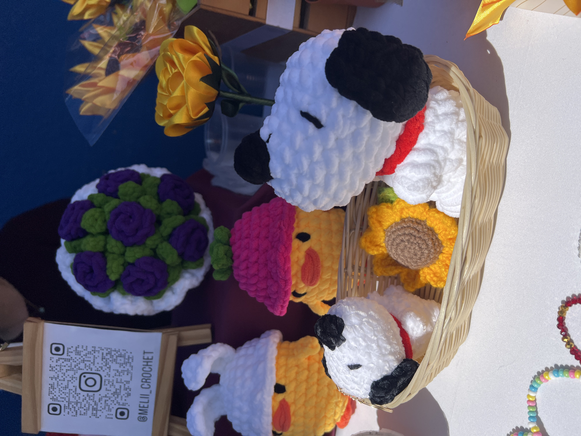 The image shows a tabletop display of crocheted items for sale. The display features two crocheted Snoopy dog heads with red collars in a small wicker basket, along with two crocheted carrot figures and a crocheted sunflower. Also on the table is a crocheted floral arrangement with purple roses and green leaves in a white crocheted base. There is a sign with a QR code and the text "@MELII_CROCHET". The scene likely takes place in Arica, Chile, although the specific location within the city is not specified.
