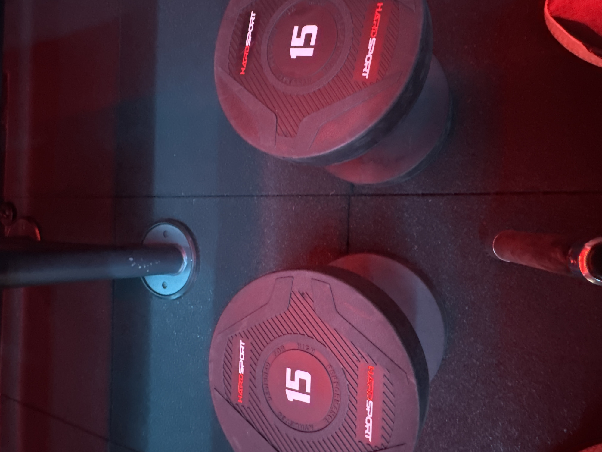 The image depicts two 15 kg dumbbells resting on a dark floor. The dumbbells are circular with the brand "HARDSPORT" clearly visible in red lettering, along with the weight. The floor appears to be a rubberized gym mat. In the background, a silver metal pole is partially visible, attached to the floor with a circular base. Red ambient lighting casts a glow over the scene, with some blue light reflecting on the floor. The image is shot from a slightly elevated angle. There are no people visible in the image. The location in Almaty, Kazakhstan, is a gym.
