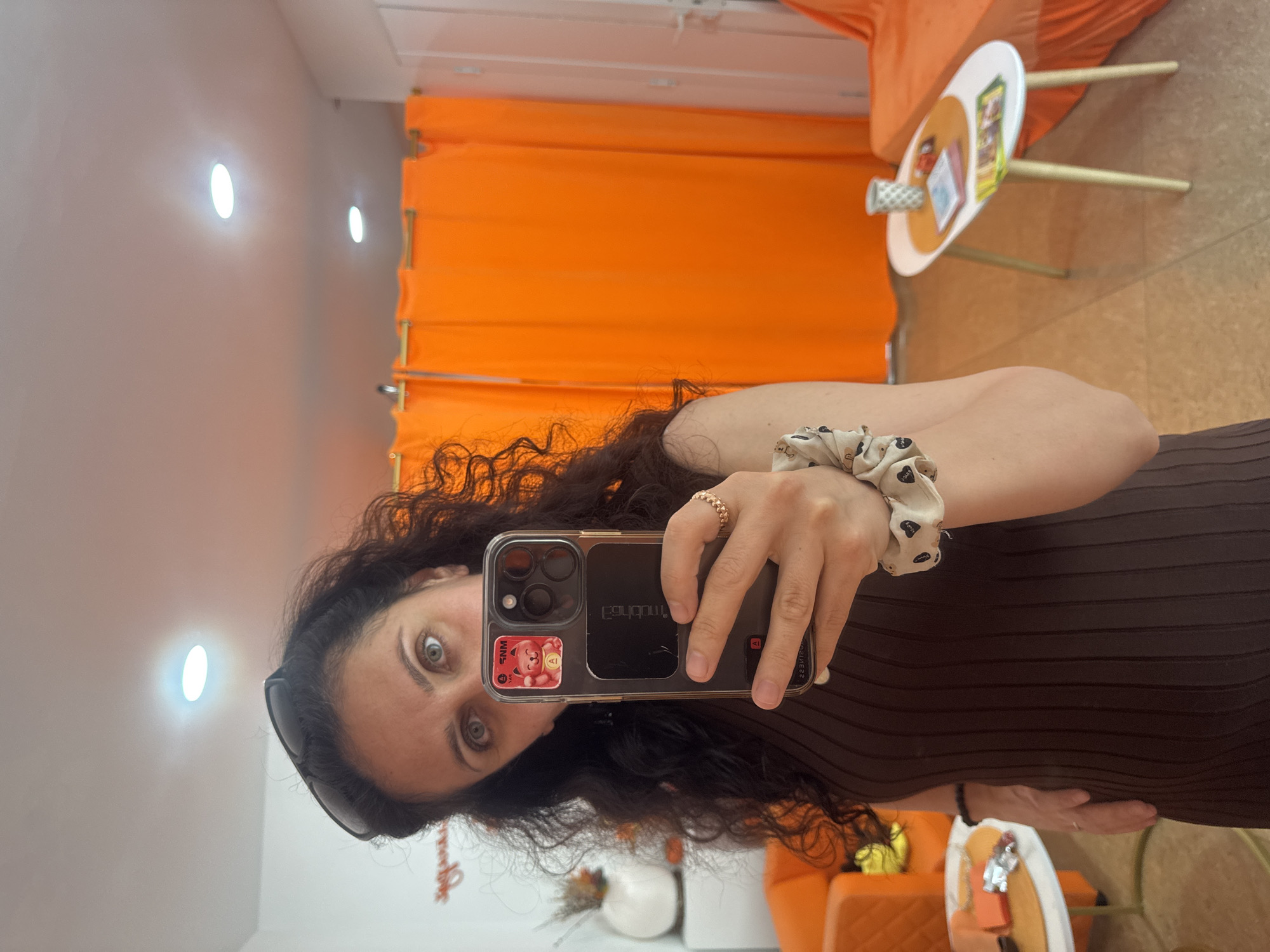 An indoor mirror selfie captures a woman with long, dark curly hair and light eyes, wearing a dark ribbed top. She holds an iPhone with a clear case, which has a sticker featuring a red bear and the text "WNS". Mirrored text on the phone case includes "gqLdnq" and "233MS2". A scrunchie is on her wrist and a ring on her finger. Sunglasses rest on her head. The background features bright orange pleated curtains or fabric panels, matching orange upholstered furniture (possibly a sofa or chair), and a small round table holding various small items like decorative tapes and cards. Two oval wall lights illuminate the light-colored wall. A white decorative pumpkin is visible on a lower shelf or surface. The scene portrays an inviting, well-lit indoor space. The location context is Sochi, Russia.