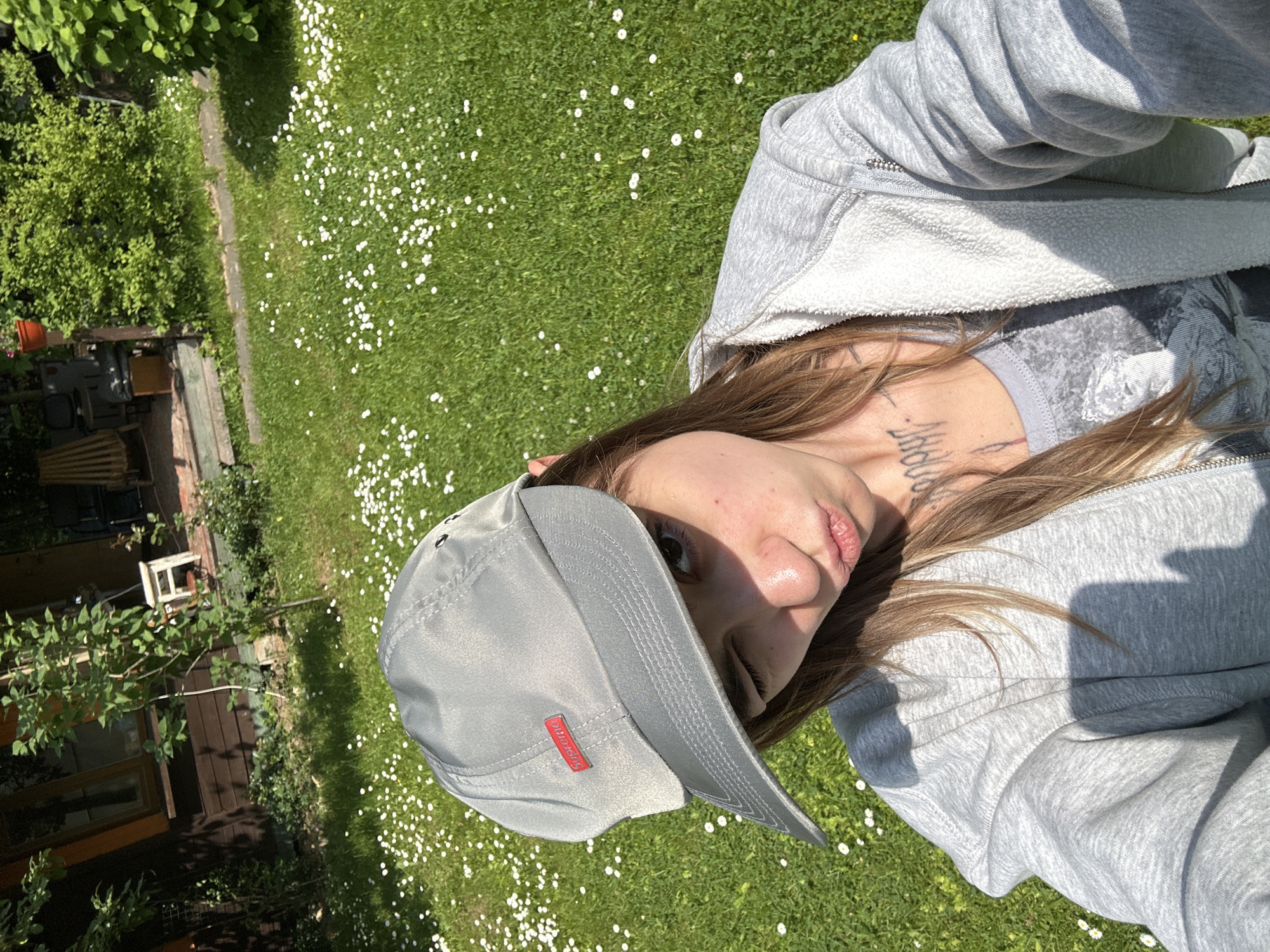 A young person wearing a grey baseball cap and a grey hoodie poses for a selfie in a grassy yard. The yard is dotted with small white flowers and a wooden building is visible in the background. The person is winking and puckering their lips. The scene appears to be in a residential area of Козино, Russia.