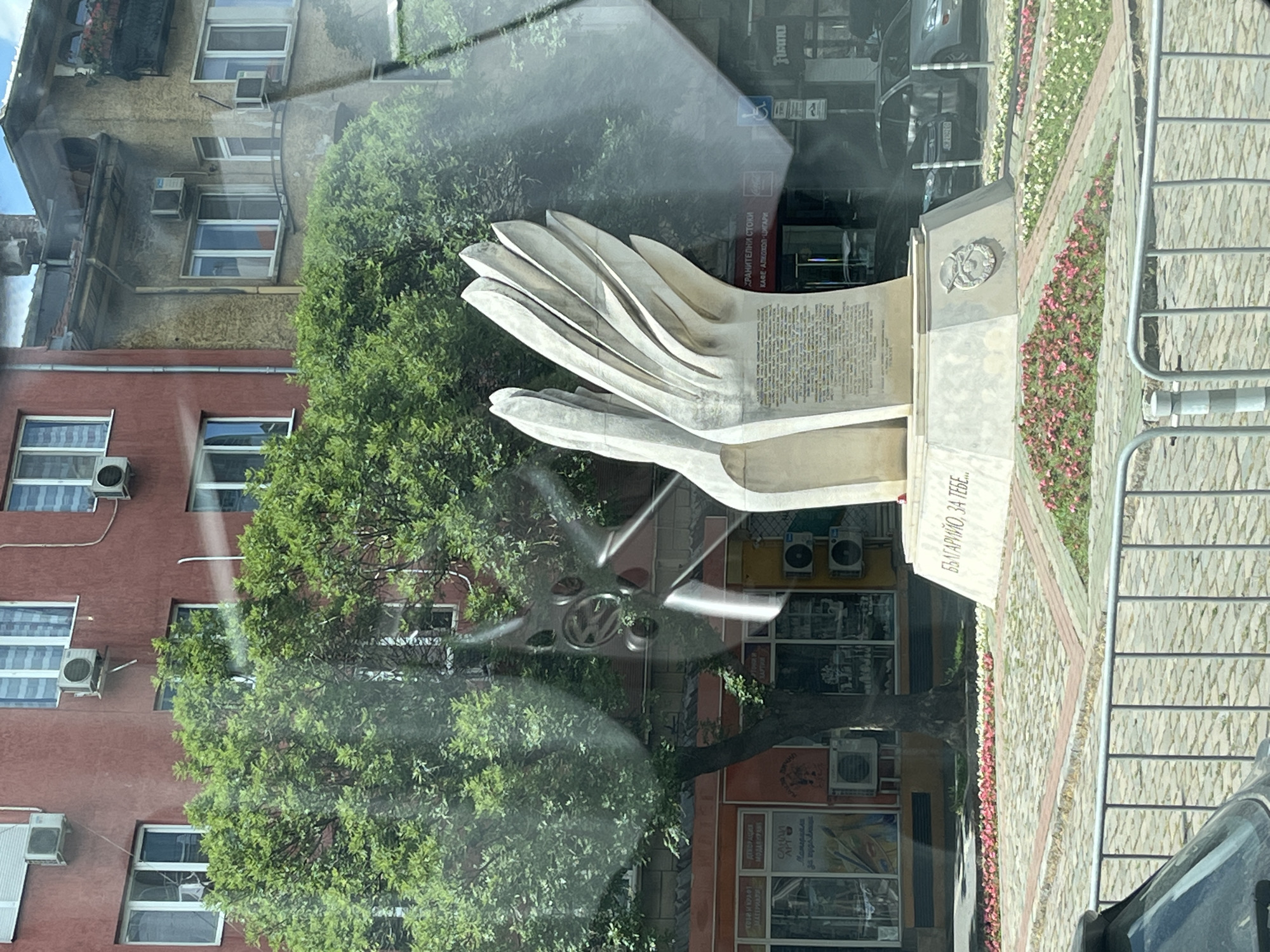 The image shows a sculpture of hands, possibly a monument, situated outdoors in what appears to be a city or town square. The sculpture is made of a light-colored material, with several curved layers forming the hands. Behind the sculpture, a building with red brick facade and numerous windows is visible, along with lush green trees. The ground in front of the sculpture is paved with stone and features flower beds with pink and white flowers.
Reflections on the surface in front of the sculpture suggest it might be viewed through a window, possibly from a vehicle. A car's wheel, featuring a Volkswagen logo, is reflected prominently in the foreground. The lighting suggests it is daytime, with the sun casting shadows and illuminating the scene.
Visible text in the image includes Cyrillic inscriptions. One prominent inscription on a stone slab reads "БЪЛГАРИЙО, ЗА ТЕБЕ..." which translates to "Bulgaria, for you...". Other text, partially obscured by reflections, appears to be on shop signs behind the sculpture, mentioning "ХРАНИТЕЛНИ СТОКИ" (Groceries) and "КАФЕ АЛКОХОЛ ЦИГАРИ" (Coffee, Alcohol, Cigarettes). The overall scene conveys a public space, likely a memorial or monument in a Bulgarian town or city.