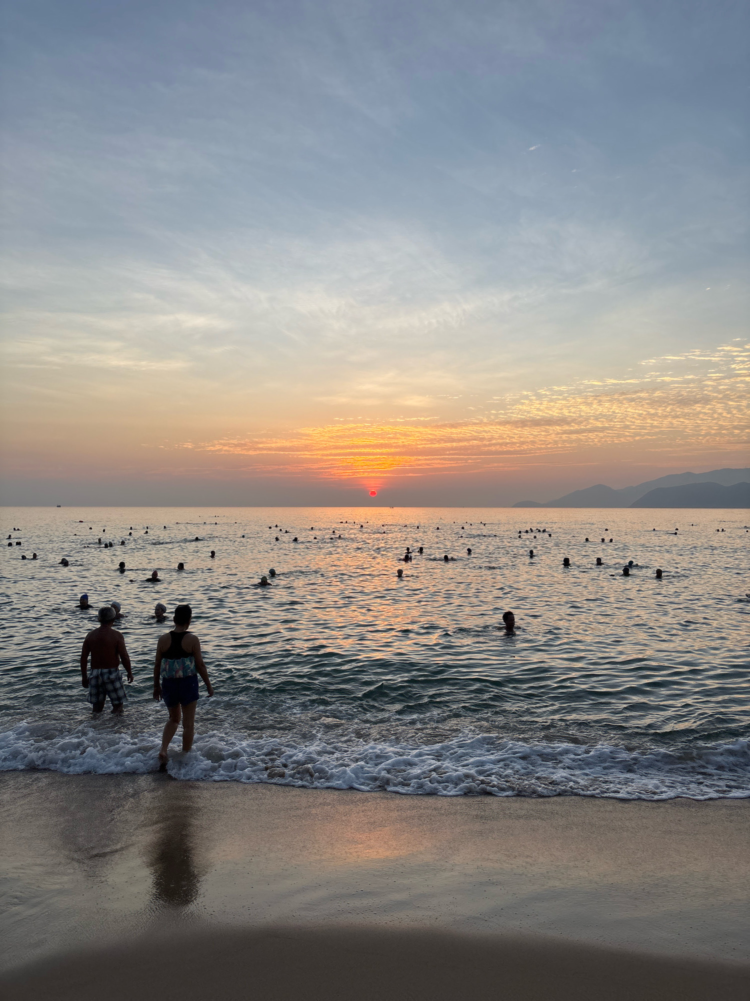 The image depicts a crowded beach scene at sunset in Nha Trang, Vietnam. The sun, a bright red orb, is low on the horizon, casting a warm orange glow across the sky and water. The sky is a gradient of blue and orange, with wispy clouds adding texture. Numerous people are visible in the water, some swimming, others standing or wading near the shore. In the foreground, two individuals, a man and a woman, are standing at the edge of the water, facing the sea. The man is wearing plaid shorts, and the woman is wearing shorts and a patterned top. Gentle waves are breaking on the sandy shore, creating white foam. In the distance, the silhouettes of mountains can be seen against the fading light. A small boat is visible on the far left of the horizon.