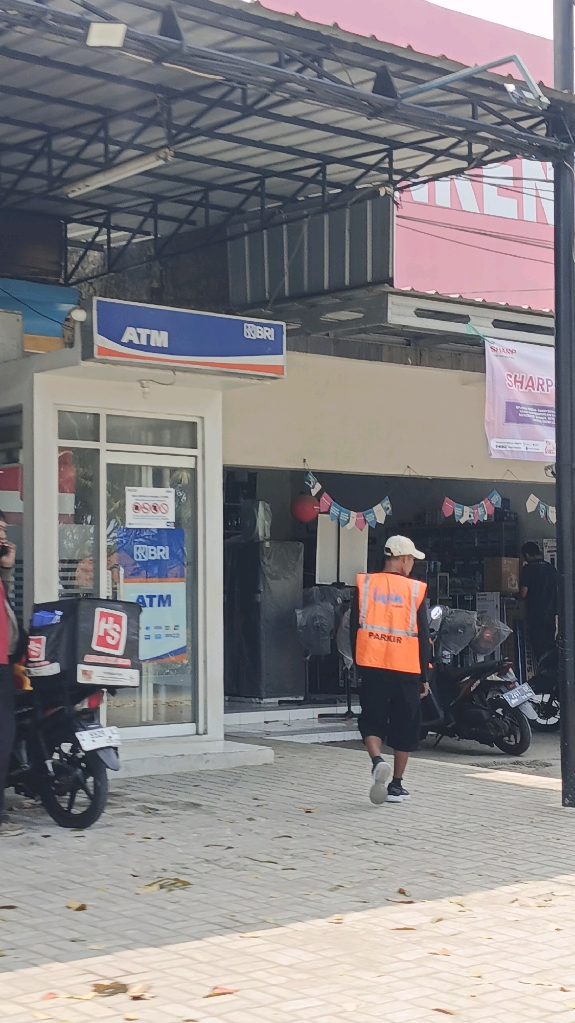 In Desa Wonoharjo, Indonesia, an ATM booth for Bank Rakyat Indonesia (BRI) is visible, featuring a blue and orange sign with "ATM" and the BRI logo. A person wearing a bright orange vest with the word "PARKIR" (parking) printed on it walks away from the ATM towards the right of the frame. The surrounding environment appears to be a commercial area with shops. A motorbike is parked to the left of the ATM, and another motorbike is parked to the right, near the person in the orange vest. The ground is paved with light-colored bricks, and fallen leaves are scattered on the pavement. In the background, shops with banners and displays are visible.