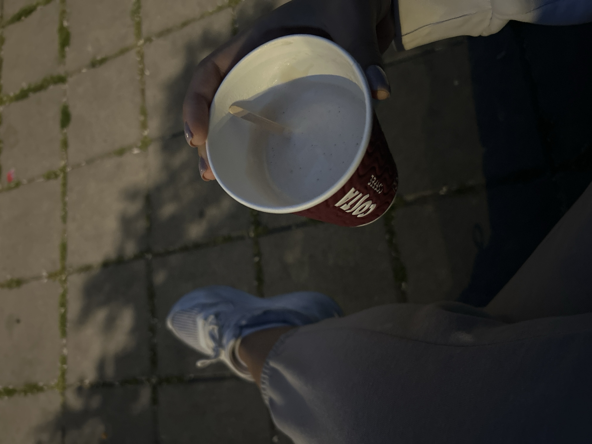A high-angle shot shows a person holding a Costa Coffee cup in their left hand. The cup contains a white beverage and a wooden stirrer. The person is wearing a light-colored jacket and grey pants. The person also has a light blue shoe on. The location appears to be outdoors on a brick paved sidewalk.