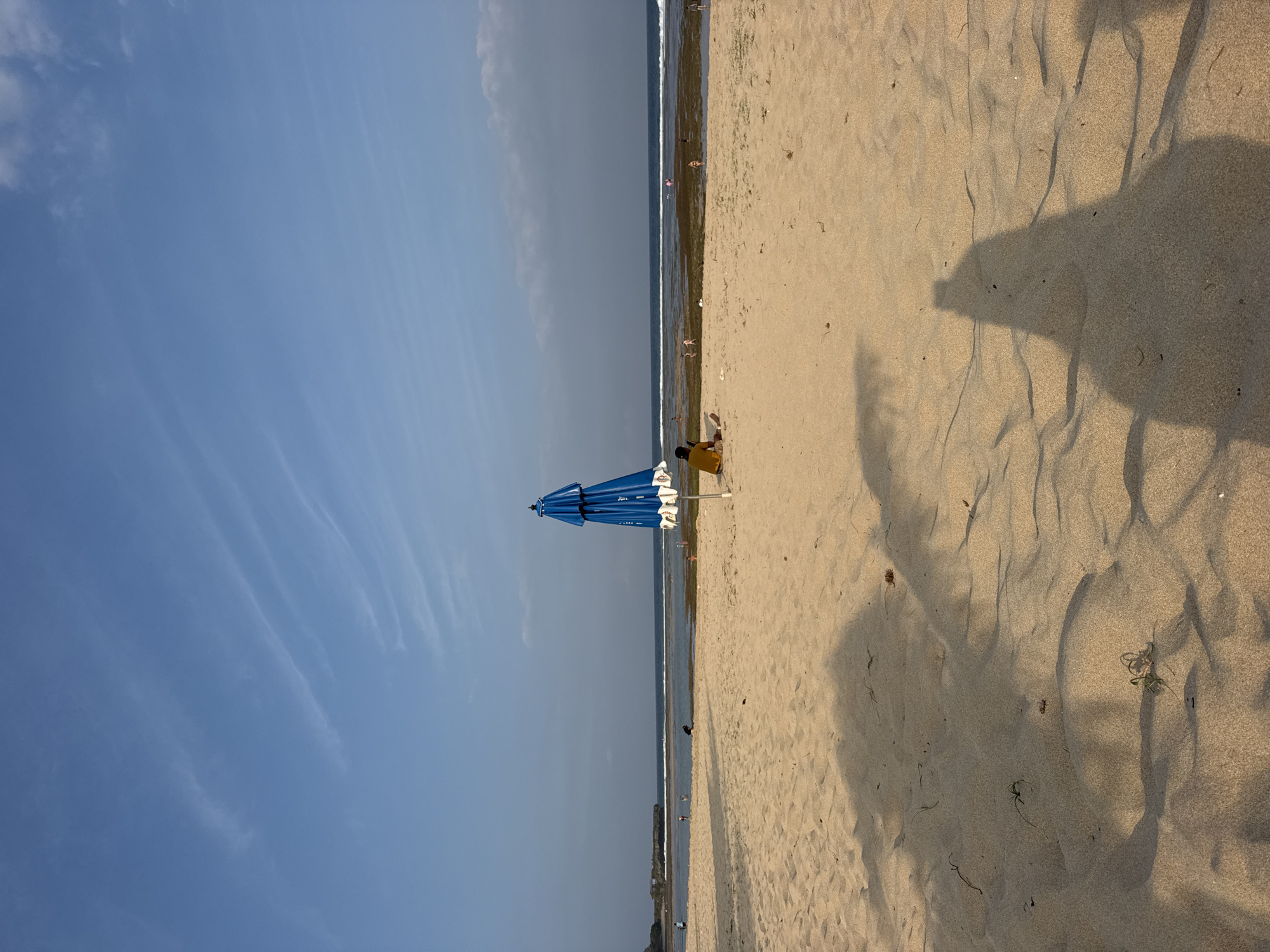The image depicts a beach scene in Kuta Selatan, Indonesia. In the foreground, sand dunes are cast in shadow. A blue beach umbrella stands in the middle ground, slightly to the left of center, with a person sitting beside it, facing away from the camera, looking out towards the ocean. The ocean is visible in the background with gentle waves. Several other people are scattered along the shoreline and in the water. The sky is a clear blue with faint, wispy clouds. The overall atmosphere is tranquil and sunny.