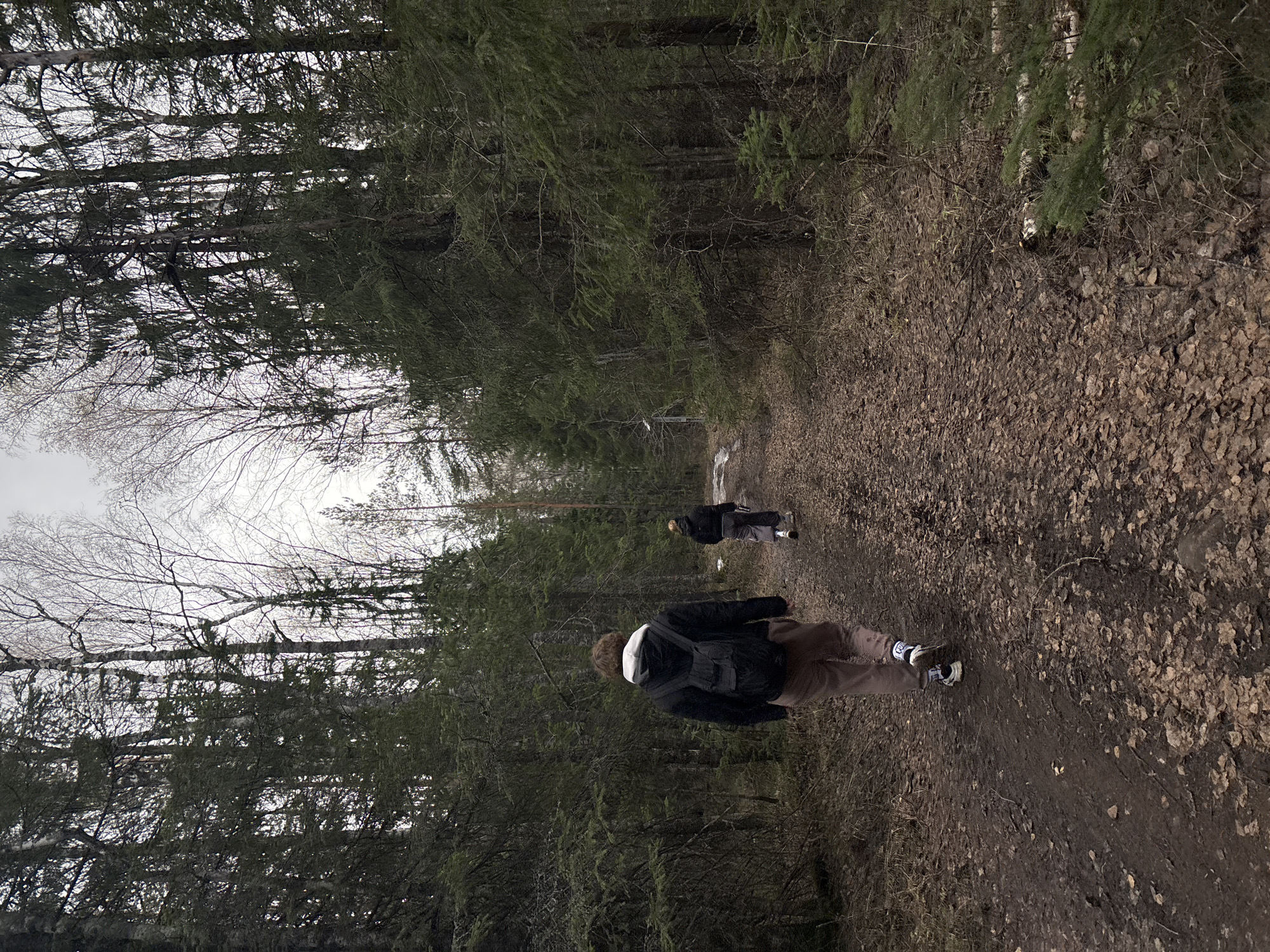 Two individuals walk along a dirt path covered in fallen leaves. The path is bordered by dense forest. The location is the city of Kachkanar, Russia. The first individual, closer to the viewer, wears a dark jacket, tan pants, and sneakers with white socks. They have a backpack on. The second individual, further down the path, is wearing darker clothing. The sky is overcast.