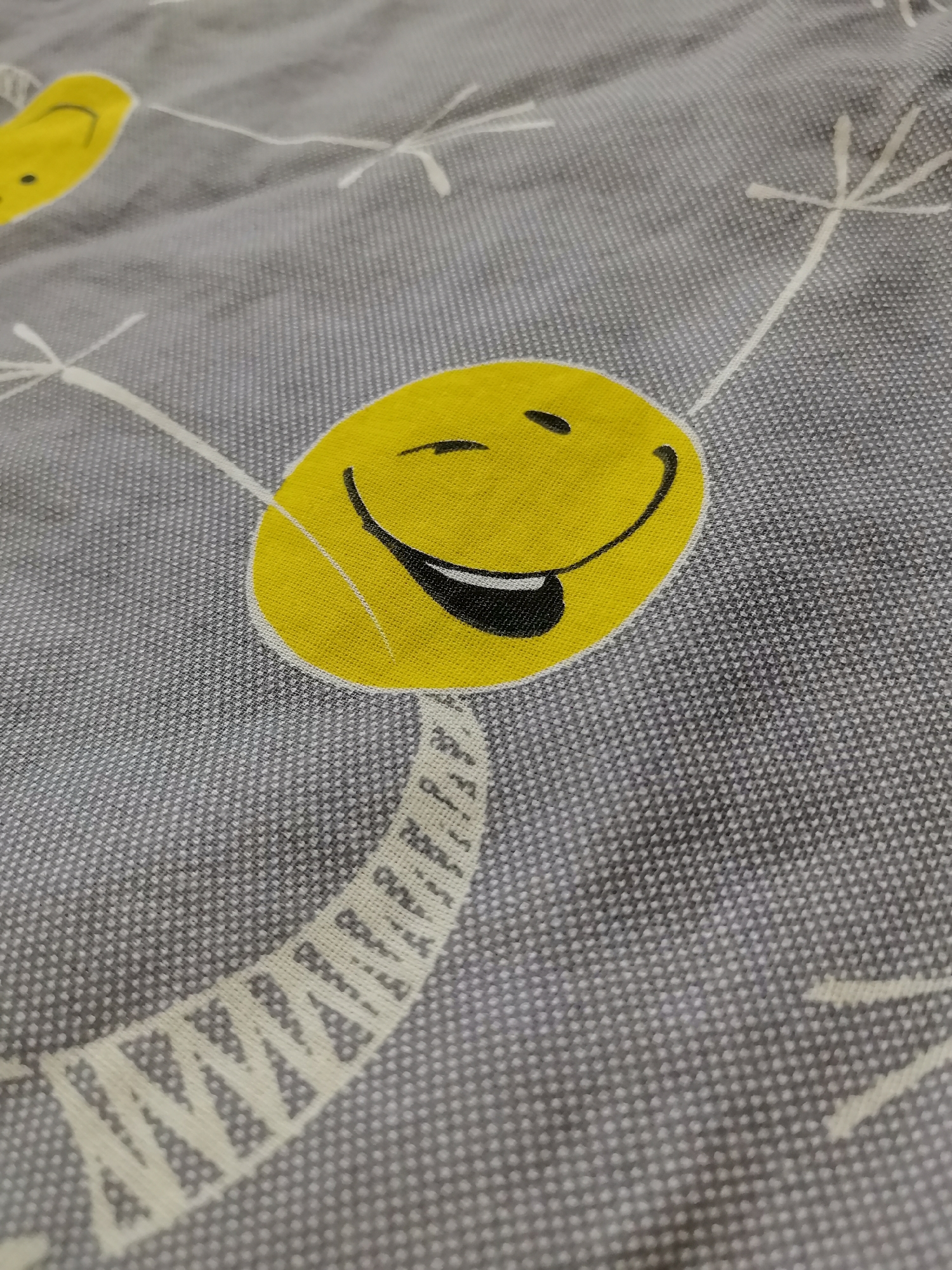 The image shows a close-up of a grey fabric with a pattern of yellow smiley faces and white dandelion-like shapes. One large, prominent smiley face is in the center right of the frame, with its mouth open and tongue sticking out. Another partial smiley face is visible in the upper left corner. The fabric has a textured appearance, likely due to its weave. There are no people or discernible elements of the environment or city of Zelenogorsk, Russia, in the image.