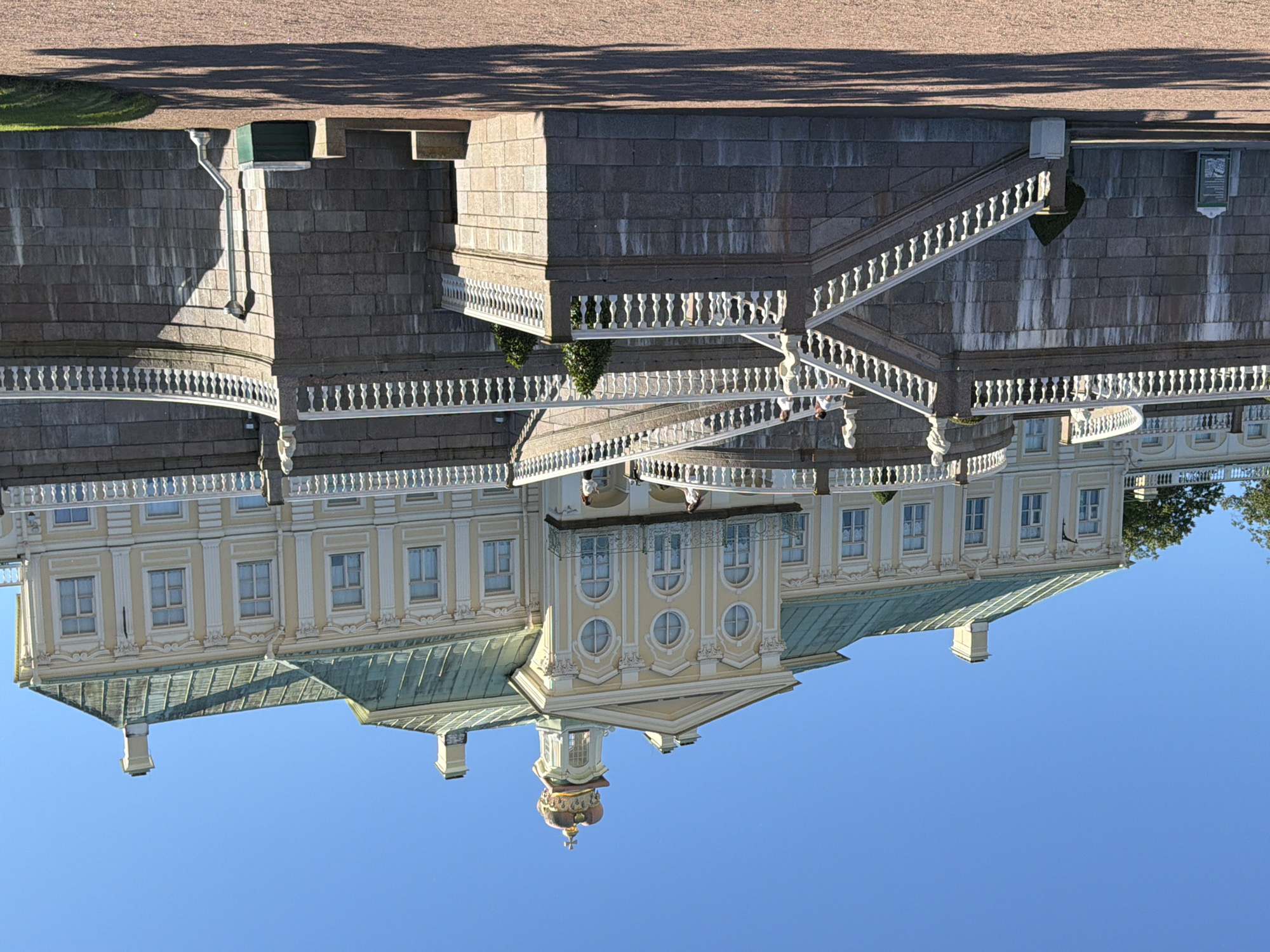 This is an eye-level, medium-wide shot of a grand, pale yellow palace with a green roof, topped by a golden crown on a central turret. The palace has numerous windows and decorative architectural elements. In front of the palace is a monumental stone staircase with white balustrades. Several people are visible on the stairs, some walking and some standing. The staircase descends to a gravel-covered area in the foreground, with shadows of trees cast upon it. The sky is clear and blue. The overall scene suggests a sunny day at a historical landmark.