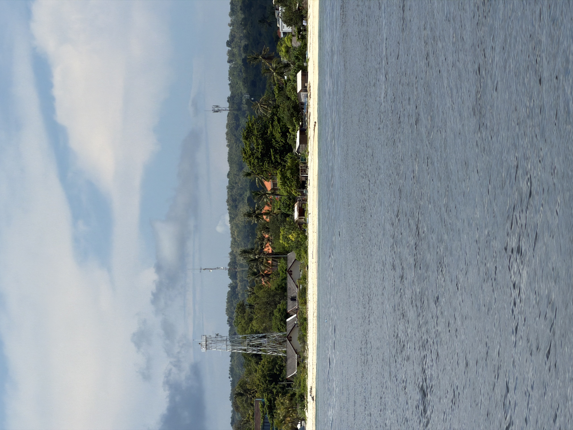 The image depicts a coastal scene in Jungutbatu, Indonesia. The view showcases a beach with light sand bordering a body of water, likely the ocean. The water is a pale blue-grey with ripples and variations in tone. On the land, a thin strip of beach is followed by a line of structures, appearing to be bungalows or houses, with dark-colored roofs. Behind these structures, dense vegetation dominates, including trees and shrubs. Interspersed within the vegetation are communication towers, tall metal structures. The sky is visible on the left side of the image, displaying a mix of pale blue and white clouds.